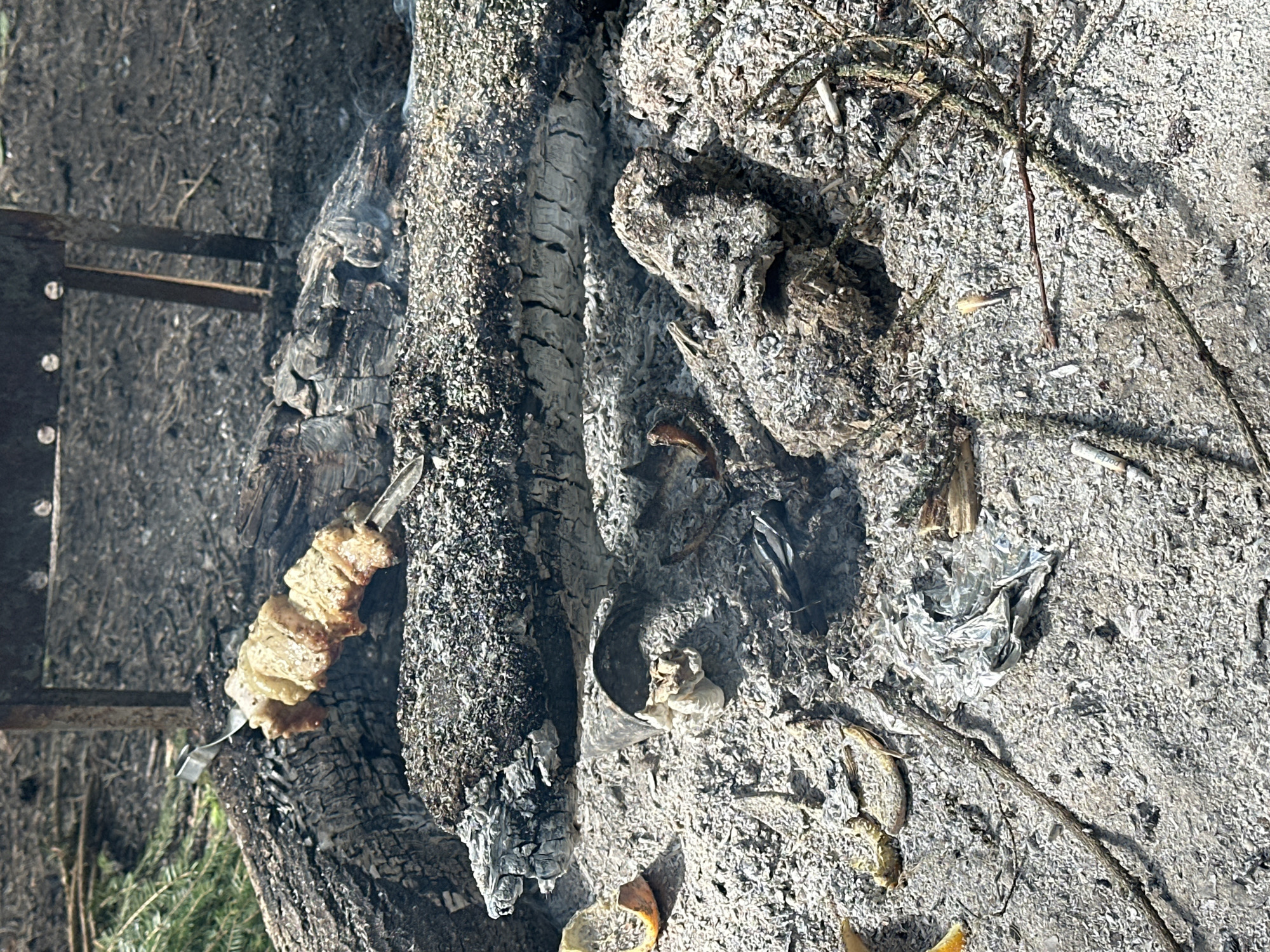 The scene depicts the aftermath of a fire. Ash and charred wood dominate the view. A metal skewer with partially cooked food rests near the burnt remains. Cigarette butts and debris are scattered among the ashes. The image suggests an outdoor setting, possibly in a rural or recreational area within or near Юзуфоўскі сельскі Савет, Belarus. No people are visible.