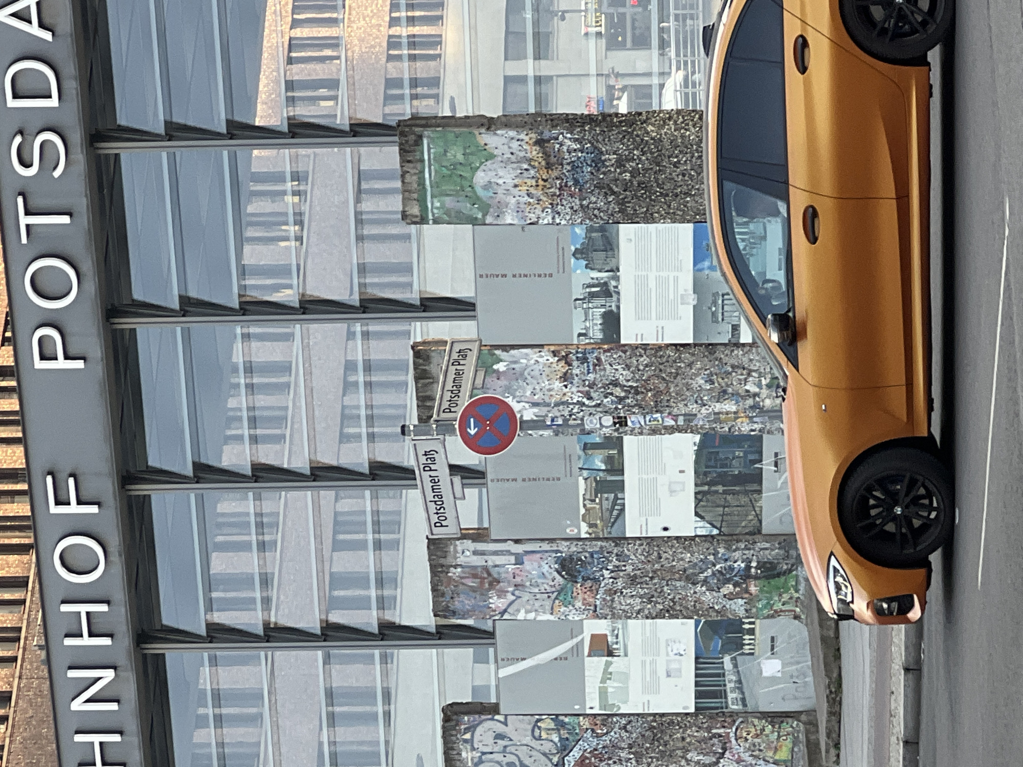 In Berlin, Germany, an orange BMW sedan is parked on the side of a street, facing right. In the background, fragments of the Berlin Wall, adorned with graffiti and informational displays, stand before a modern building with a glass facade. A signpost reads "Potsdamer Platz". The overall scene depicts a historical landmark contrasted with contemporary urban architecture and a modern vehicle.