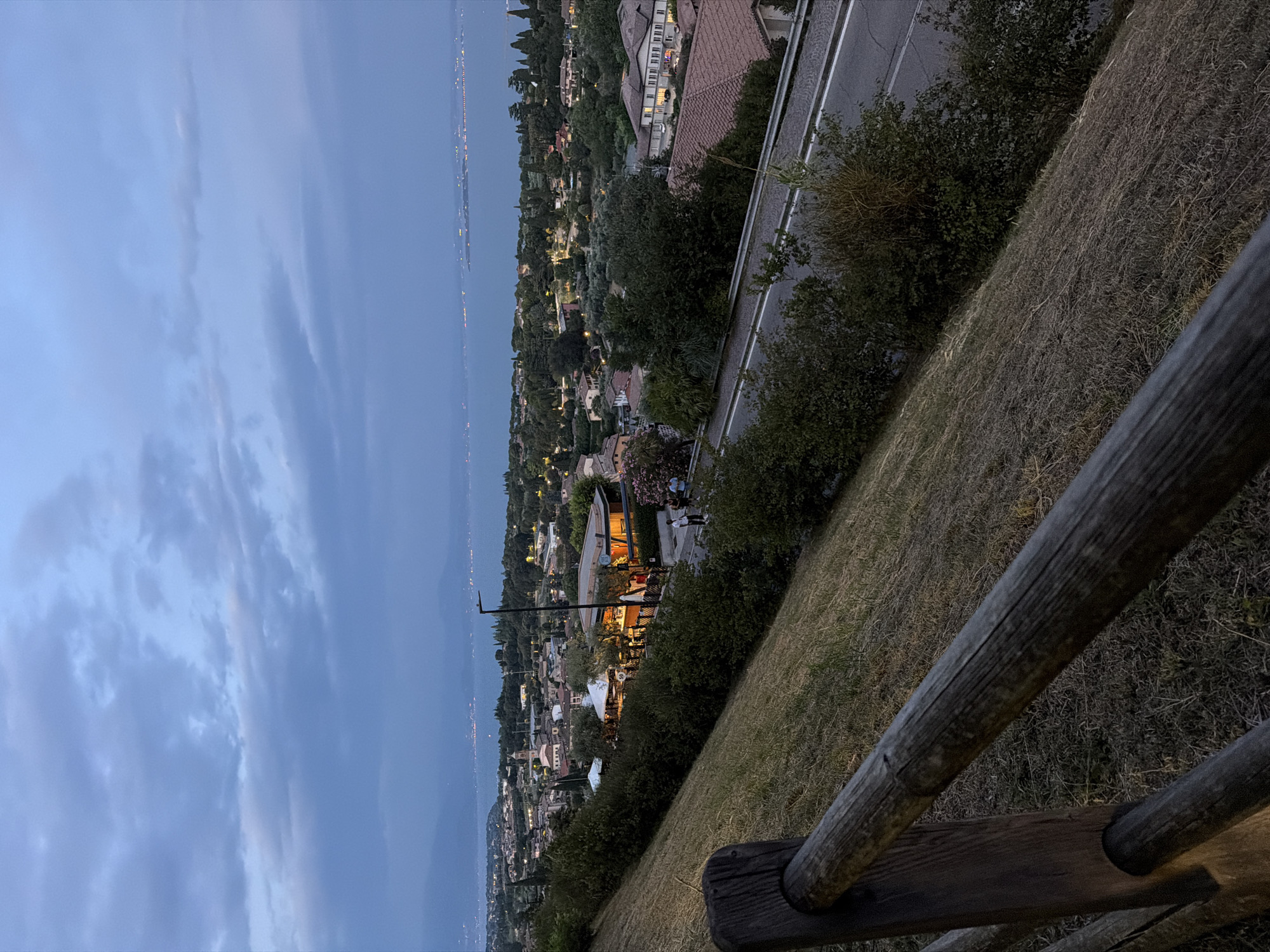 The image depicts a twilight view overlooking Padenghe sul Garda, Italy, from an elevated vantage point. In the foreground, a rustic wooden fence runs diagonally across the frame, with dry, mowed grass covering a steep embankment. Beyond the fence, a road curves through a landscape of buildings and dense greenery.
The town of Padenghe sul Garda is visible in the midground, characterized by its red-tiled roofs and scattered lights, indicating the onset of evening. A prominent building with illuminated windows and an orange glow stands out amongst the trees. A few people are seen walking on a paved area near this building and along the road. The town extends towards a large body of water, likely Lake Garda, which dominates the background. The lake reflects the dimming sky, and lights from distant towns or structures are visible along its shoreline, creating a hazy, luminous line. The sky itself is overcast with large, textured clouds in shades of blue and gray, suggesting either dusk or an approaching weather change. The overall atmosphere is serene and twilight-like.