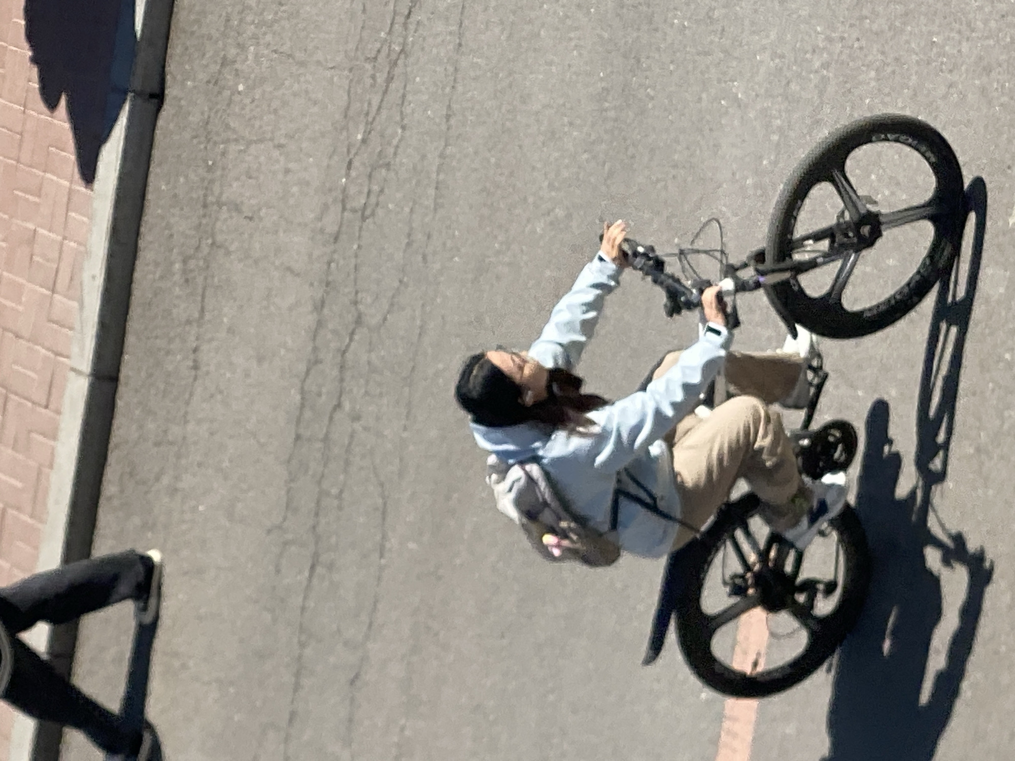 An overhead shot shows a person riding a bicycle on a gray asphalt surface in what appears to be an urban area. The person is wearing light-colored clothing, beige pants, and sneakers. They are also wearing a backpack and riding on the bicycle with their arms raised, appearing to be performing a wheelie. The bicycle has three-spoke wheels. The lower left corner shows part of a person wearing black pants and white sneakers. Part of a brick sidewalk can be seen on the left side of the image. This image was not taken in Zhongtun, China.