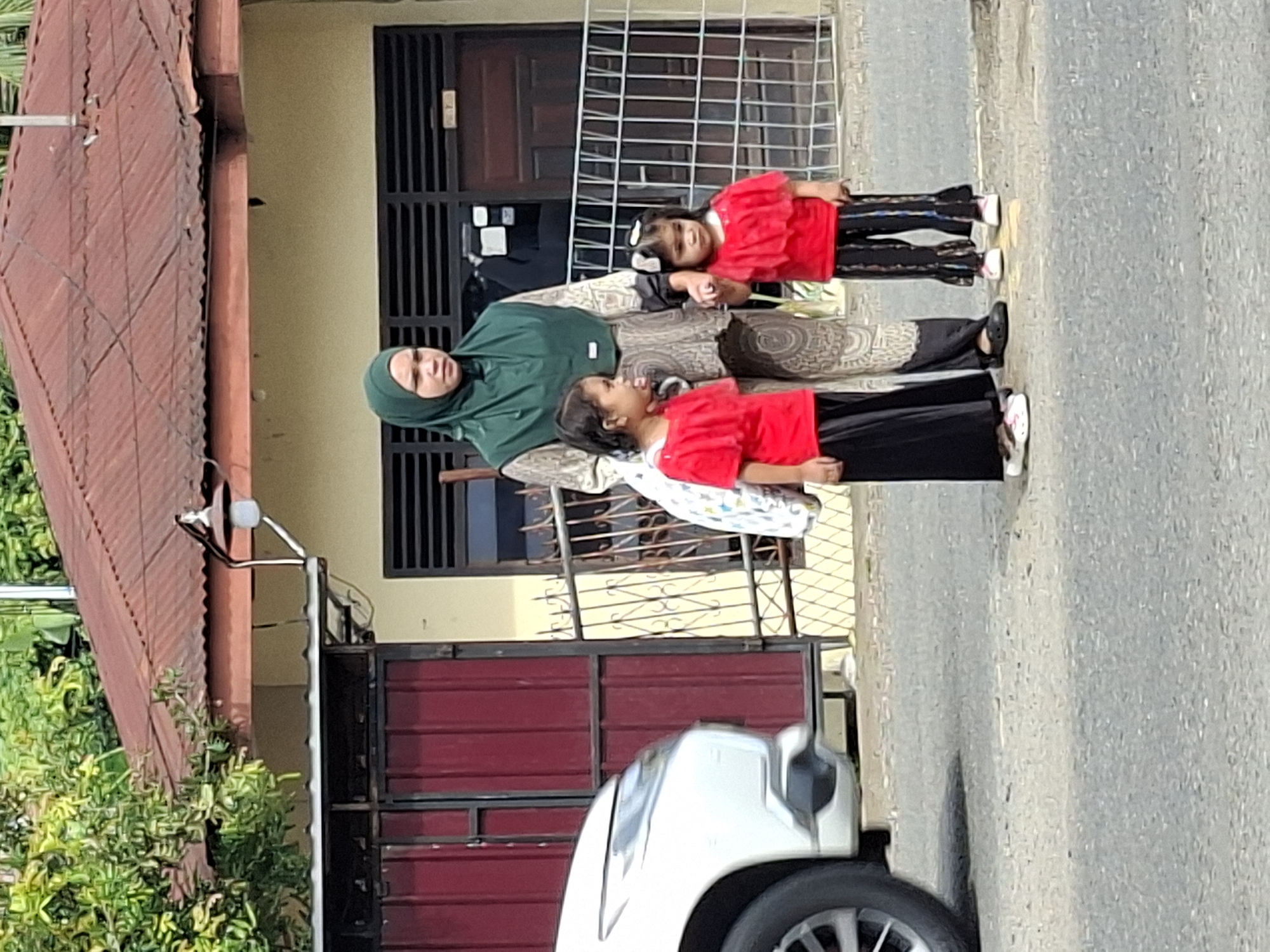 In Lhokseumawe, Indonesia, a woman wearing a green hijab stands with two young girls in front of a building with a corrugated metal roof and a light-colored facade. The woman is wearing a patterned top and pants, and the girls are both dressed in red tops and dark pants. The girl on the left wears black pants, and the girl on the right wears black pants with a colorful pattern. Both girls are holding hands with the woman. The scene appears to be outdoors, with trees visible in the background. A white vehicle is partially visible on the left side of the image. A metal fence is behind the group.
