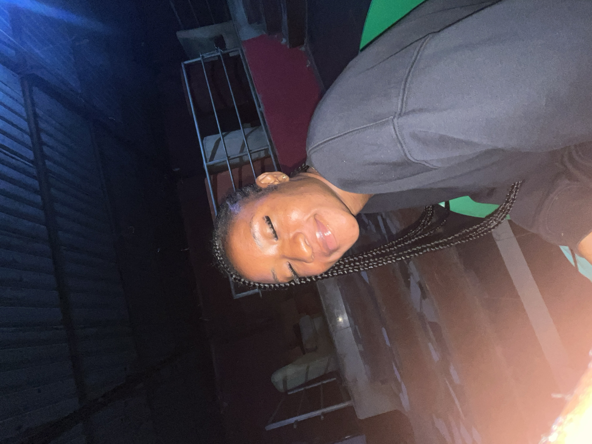 A young woman with braided hair smiles with her eyes closed, her face illuminated by a warm light. She wears a dark sweatshirt. The background suggests an outdoor setting at night, possibly a rooftop or elevated area with a corrugated metal roof overhead. In the distance, there are visible stairs and railings, indicating a multi-level structure. The overall scene appears to be in Gura Top, Nigeria, with the ambient lighting and architectural elements consistent with the region.