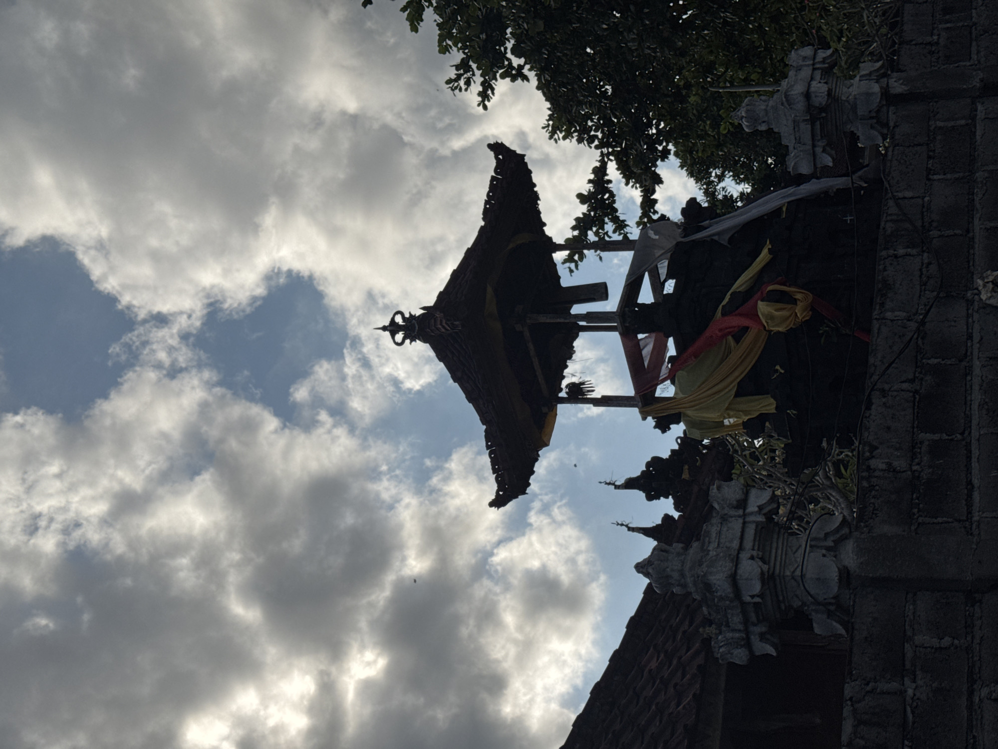 The image captures a traditional Balinese architectural structure, likely a temple or part of a temple complex, silhouetted against a cloudy sky. The scene is observed from a low angle, looking upwards. Prominently featured is an ornate, multi-tiered dark roof, possibly made of thatch or dark wood, with intricate decorative elements and a distinct finial at its apex. Lush green foliage from trees partially frames the top of the structure on the right side.
Below the roofline, parts of the building are adorned with vibrant red and yellow ceremonial sashes or fabrics, suggesting traditional or religious significance. To the right, dark stone walls, possibly cinder blocks, are visible, alongside carved stone statuettes or decorative elements that appear to be part of the building's facade. The sky above is largely covered with white and grey clouds, though patches of clear blue sky are also visible. The lighting suggests an overcast day or possibly late afternoon, creating a strong silhouette effect for the building against the brighter sky. No people or specific activities are visible, presenting a serene and static architectural landscape. There is no visible text. The setting is consistent with the architectural style found in Kuta Selatan, Indonesia.