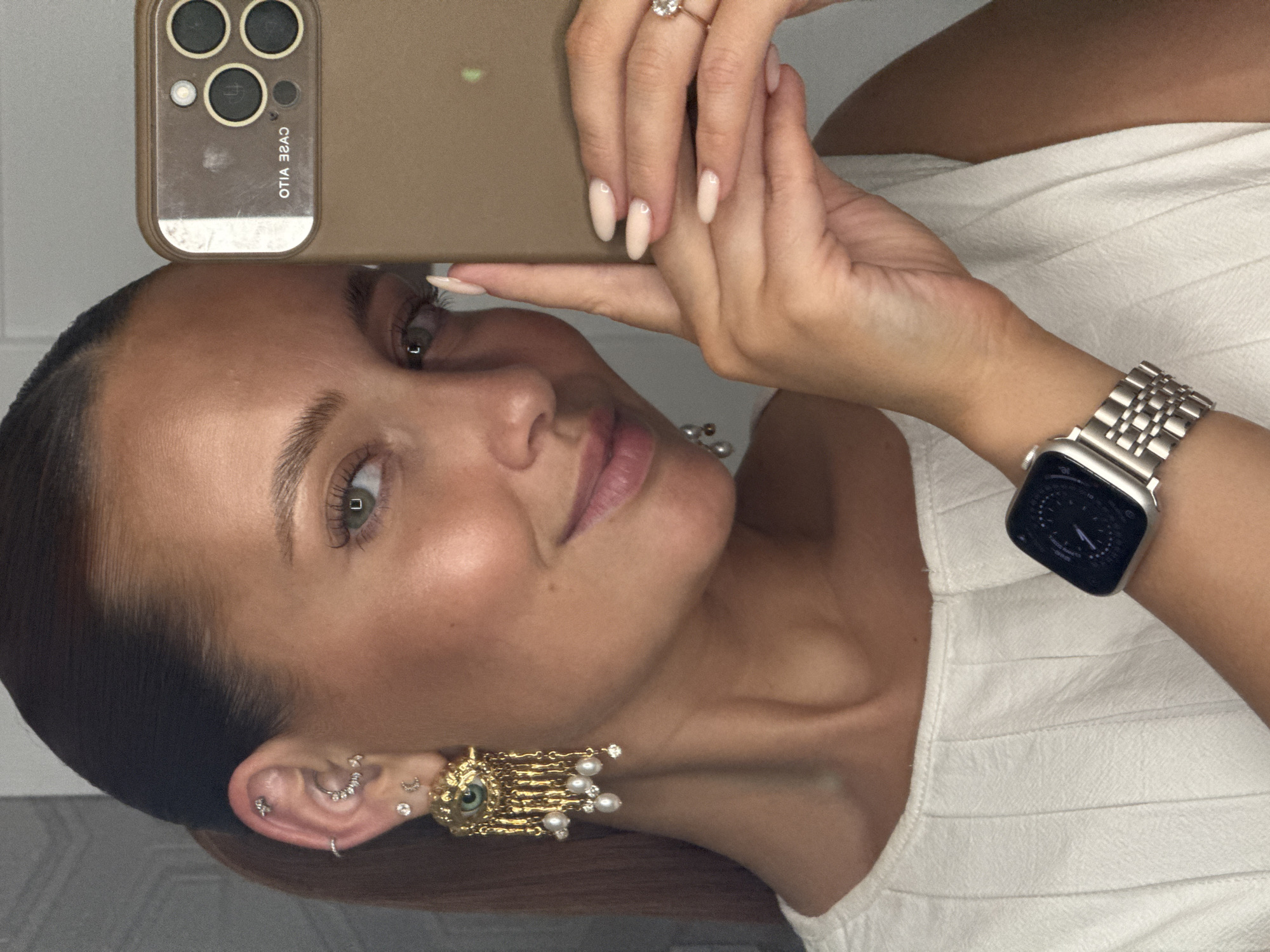 A woman is taking a selfie. She is holding a smartphone up to her face. The smartphone has a brown case with three camera lenses. She is wearing a white top and a silver watch on her left wrist. She has a ring on her finger and her nails are manicured. She is wearing a large, gold earring in her left ear, along with several other smaller earrings. Her hair is pulled back in a sleek style.
There is no information available to establish the location of the photograph in Chaikovsky, Russia.