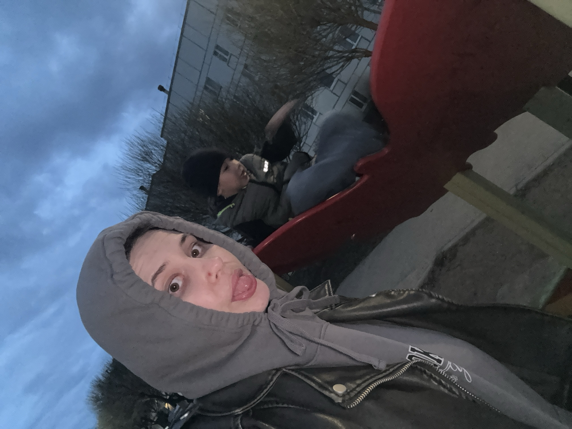Two individuals are on a playground slide in what appears to be Chaikovsky, Russia. One person, wearing a black beanie, is sitting on the slide. The second person, wearing a gray hoodie and black leather jacket, is positioned in front, facing the camera. The person in the hoodie has stuck their tongue out. A multi-story building can be seen in the background, behind leafless trees, under an overcast sky.
