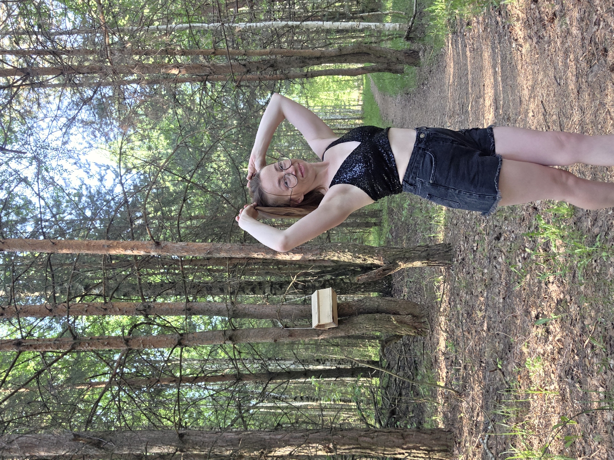 A woman stands in a forest in the city of Chaikovsky, Russia. She has fair skin, brown hair, and wears glasses. She is wearing a black sequined top and black denim shorts. Her arms are raised above her head. Behind her, a narrow path extends into the distance between the trees. A small wooden birdhouse is attached to one of the trees. The ground is covered in fallen leaves and green vegetation. The trees are tall and dense, creating a canopy overhead with glimpses of the sky.