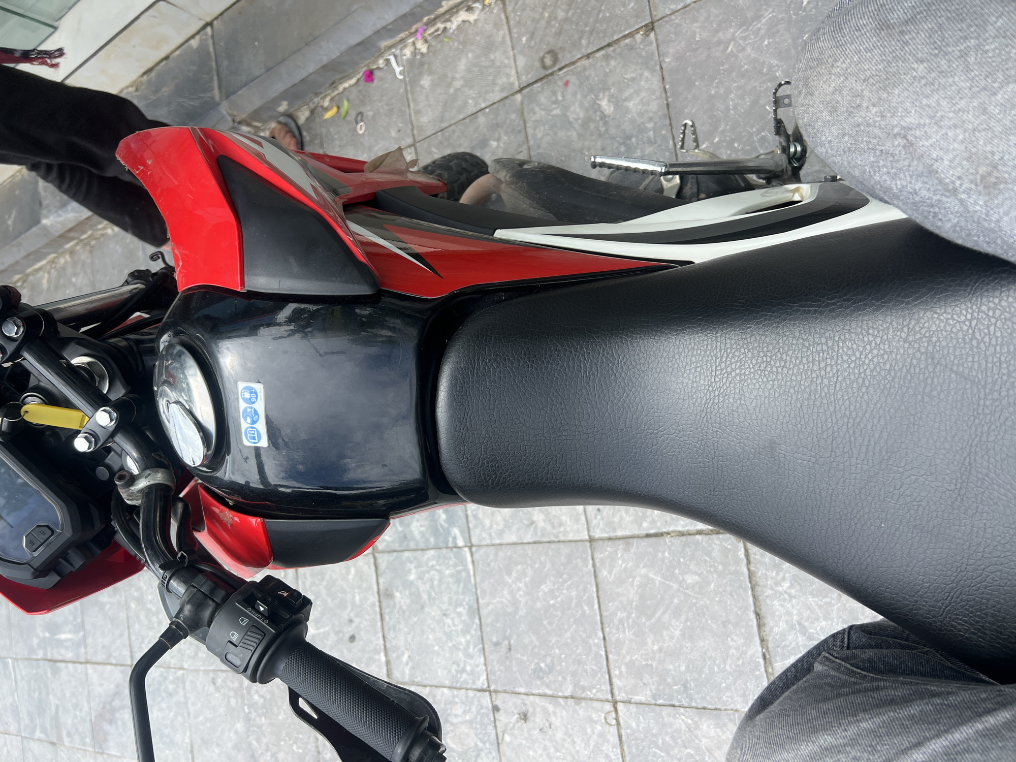 The image captures a close-up, high-angle perspective of a red and black motorcycle parked on a paved sidewalk. The motorcycle's seat is black and textured. On the fuel tank, two stickers are visible: one with a blue circular icon and another with a white circular icon featuring a blue motorcycle and the number "90+". The handlebars, with a black grip and control switches, are prominent in the upper left. Part of a person's legs, clad in grey jeans, is visible in the foreground on the left and right. The background shows a person in dark clothing standing behind the motorcycle. The sidewalk is composed of grey square tiles. The setting is the city of Hà Giang 2 Ward, Vietnam.