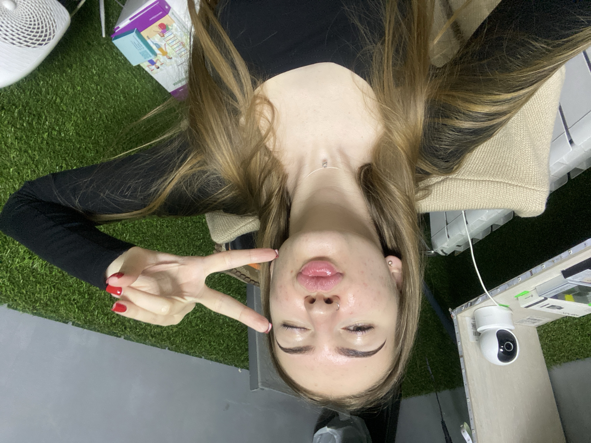 An upside-down, overhead portrait captures a young woman making a playful expression. She has long, light brown hair parted in the middle and is wearing a black long-sleeved top with a light beige knitted sweater or cardigan. Her right hand, adorned with red nail polish (some tips are white), is raised to form a peace sign. Her eyes are closed, and her lips are puckered. She has a septum piercing and a small stud earring in her left ear, as well as a thin silver necklace with a tiny pendant.
The background is predominantly covered with artificial green turf. In the upper left, a white, circular object with a patterned grille, possibly a fan or air purifier, is visible. A partially obscured white and purple box, featuring illustrations of children playing, rests on the turf near her head. To the lower right, a white spherical security camera with a black lens is mounted on a light-colored surface, which also has some white strip details. A plain gray surface occupies the bottom foreground of the image. The time of day and weather are not discernible due to the indoor setting. No significant text is clearly visible.