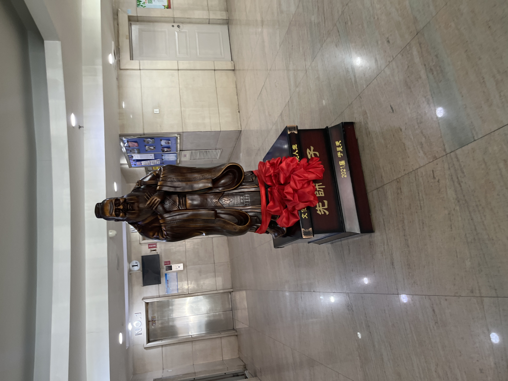 A bronze statue of a man in traditional Chinese clothing stands on a tiered, book-shaped pedestal adorned with a large red flower. The statue and pedestal are located indoors on a light-colored tiled floor, possibly marble or a similar material. In the background, there is a series of doors, a display case with information, and signage indicating an elevator. The inscription on the pedestal includes Chinese characters. The text includes the characters “先師” which translates to “Exemplary Teacher” and the characters “2021届 李昊天” which translates to “2021 Session Li Haotian”. The location appears to be an interior space in Changchun City, China, based on the language used.