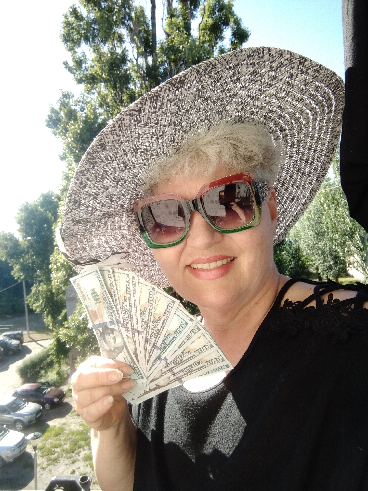 A woman wearing a wide-brimmed hat and oversized sunglasses, holding a fan of US hundred-dollar bills, smiles at the camera. She is dressed in a black top with floral embellishments on the shoulders. The background appears to be an outdoor setting with trees and a clear blue sky. Below her, a view of parked cars and a street is visible, suggesting an urban or suburban environment. The image was taken in the city of Horishni Plavni, Ukraine.