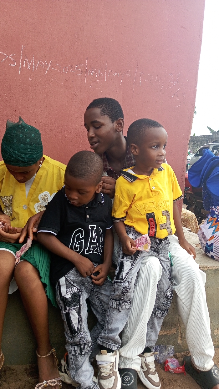 A group of four individuals, including an adult woman, an older male child or teenager, and two younger male children, are seated outdoors against a reddish-brown wall in Agburu Uke, Nigeria. The lighting suggests daylight, possibly under overcast conditions.
On the far left, an adult woman in a green headwrap and a yellow top holds what appears to be Nigerian Naira notes in her hands. Next to her, a younger boy wearing a black "GAP" t-shirt and grey patterned jeans looks down. Behind these two, an older male child or teenager in a plaid shirt is looking at the boy in black. To the right, another younger boy, wearing a yellow "VERSACE" t-shirt and similar patterned grey jeans, holds money and looks towards the right side of the frame. The patterned jeans feature various designs, including text that appears to be "GRAEL" on one of the cargo pockets. One of the children is wearing brown and white sneakers, while another has dark sandals with a "GUCCI" logo visible.
The red wall behind them has chalk or paint markings. The visible text reads "S/MAY-2025/NGA/days/T2436". In the far background, parts of a vehicle are visible. The individuals appear calm and focused, with a general sense of quiet activity, possibly related to the money being held.