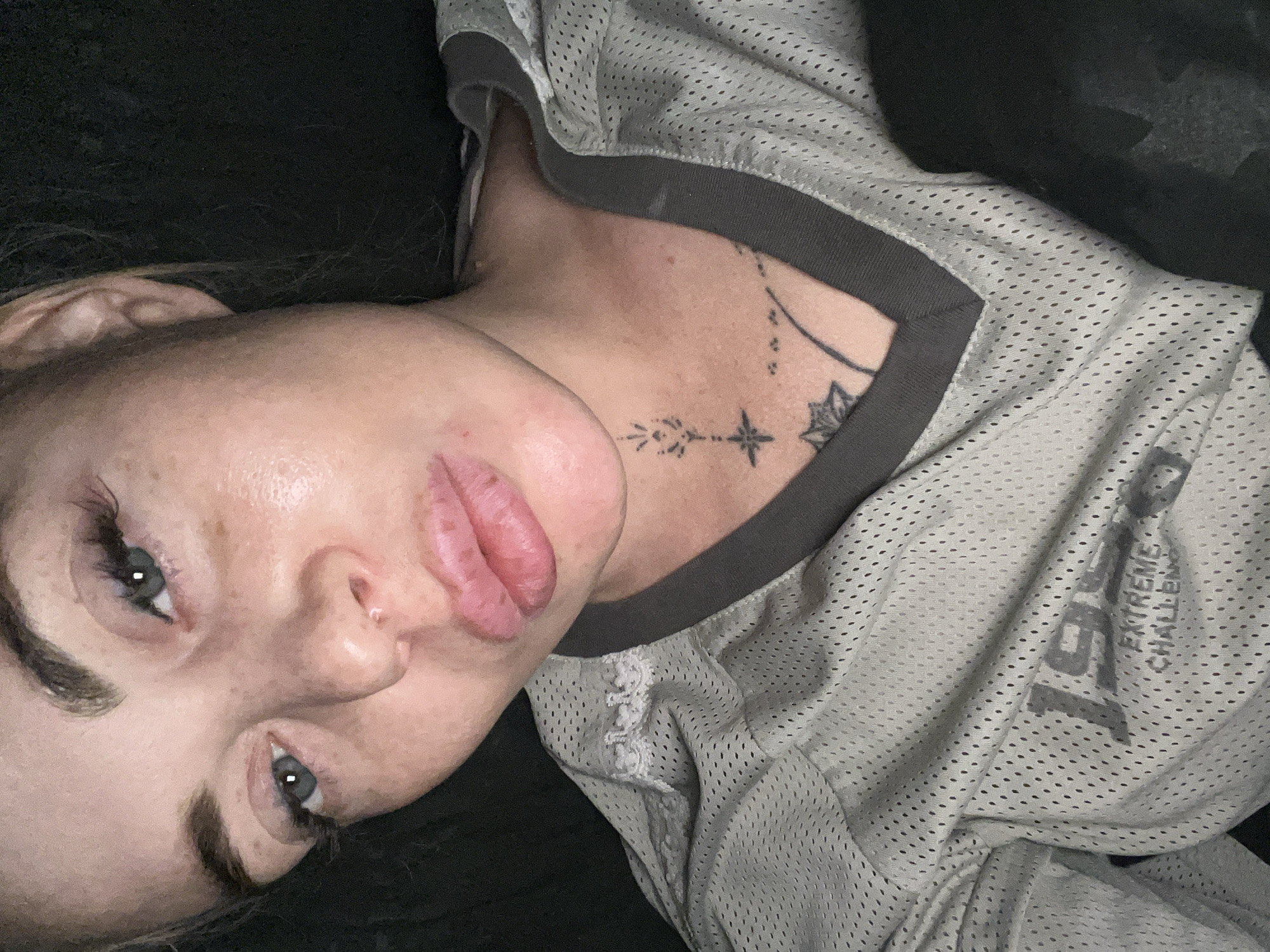 A close-up, high-angle, indoor selfie shows a woman with blonde hair pulled back, her face is centered in the frame. She has light blue eyes, full lips, and visible freckles and blemishes on her skin. Her eyebrows are dark and shaped, and she is wearing false eyelashes. She has several tattoos on her neck and chest, including a geometric design and a lotus flower. She is wearing a light gray mesh athletic jersey with a dark V-neck collar. The jersey has text on the front that reads "19XX EXTREME CHALLENGING". The background is dark and out of focus, suggesting an indoor setting with low lighting.