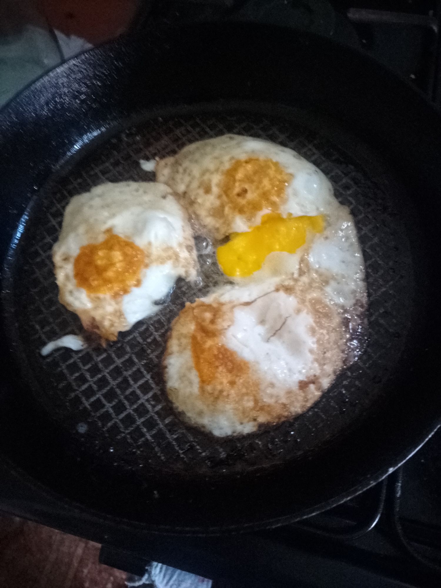 A dark, non-stick frying pan contains three fried eggs cooking on a textured grid surface. The eggs are mostly white with some browning on the edges, and one has a visible yellow yolk. The pan is set on a stovetop, with a portion of a wooden surface and a white cloth visible in the lower left corner. The setting appears to be domestic. There is no specific information that can place this image in the city of Ustyvytsia, Ukraine.
