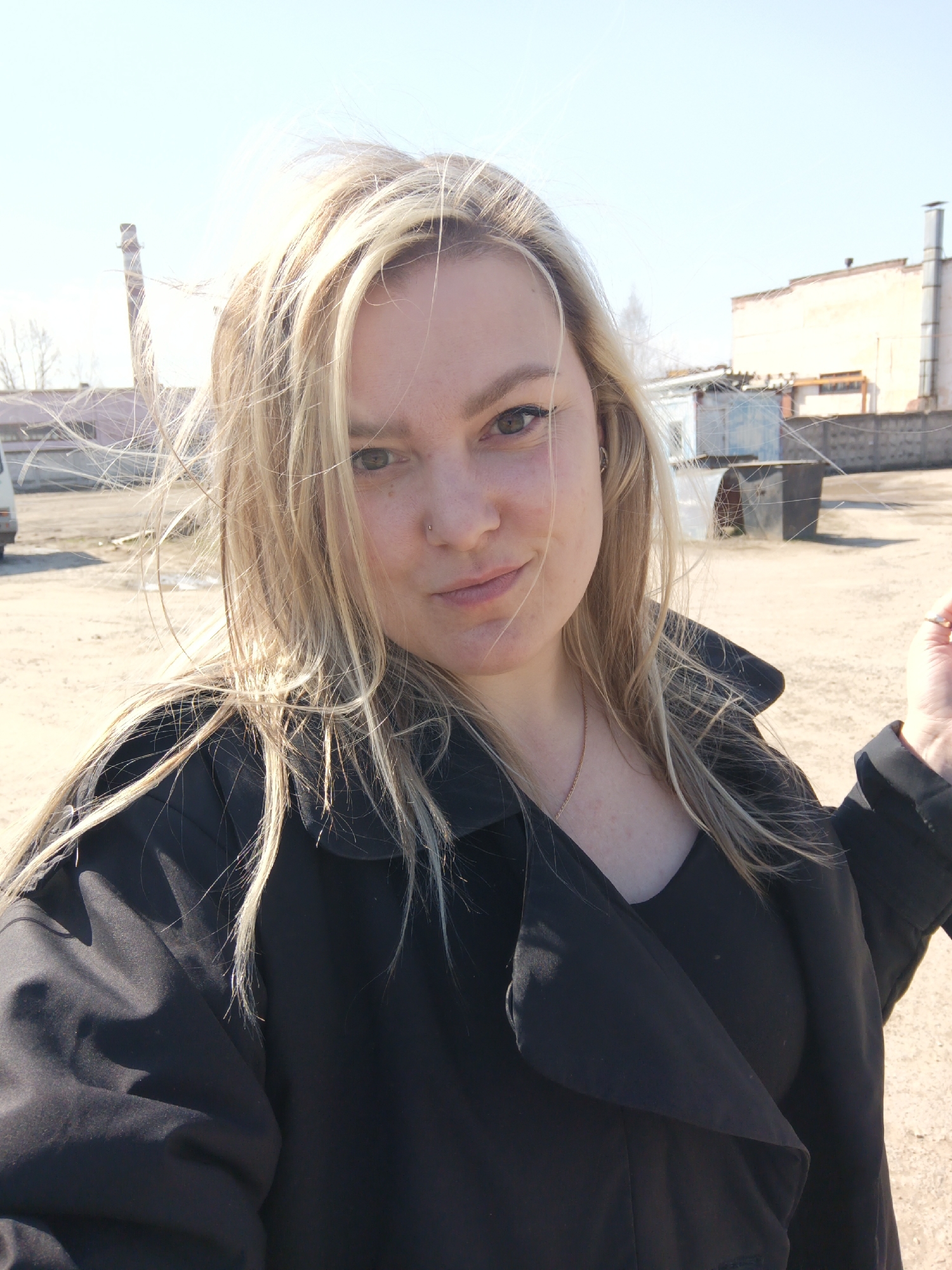 A fair-skinned woman, likely in her 20s or 30s, is captured in a front-facing selfie or close-up portrait outdoors. Her long, blonde hair is noticeably wind-blown, with strands flying across her face. She has dark, possibly green or hazel eyes and a small piercing on her left nostril. She is looking directly at the camera with a calm, subtle smile.
She is dressed in a black, collared coat or trench coat over a black top, and a thin gold chain necklace is visible around her neck. A ring with a clear stone can be seen on a finger of her left hand, which is slightly raised towards the right edge of the frame.
The background consists of an open, unpaved or dirt lot under a bright, clear blue sky. In the mid-distance, industrial or semi-industrial structures are visible, including a prominent chimney or smokestack on the left and a larger, light-colored building on the right. There are also smaller, possibly dilapidated sheds and what appears to be a concrete fence. The strong sunlight and wind-blown hair indicate a bright and breezy day. The setting suggests an industrial area, consistent with a location like Noviy, Russia. No discernible text is visible in the image.