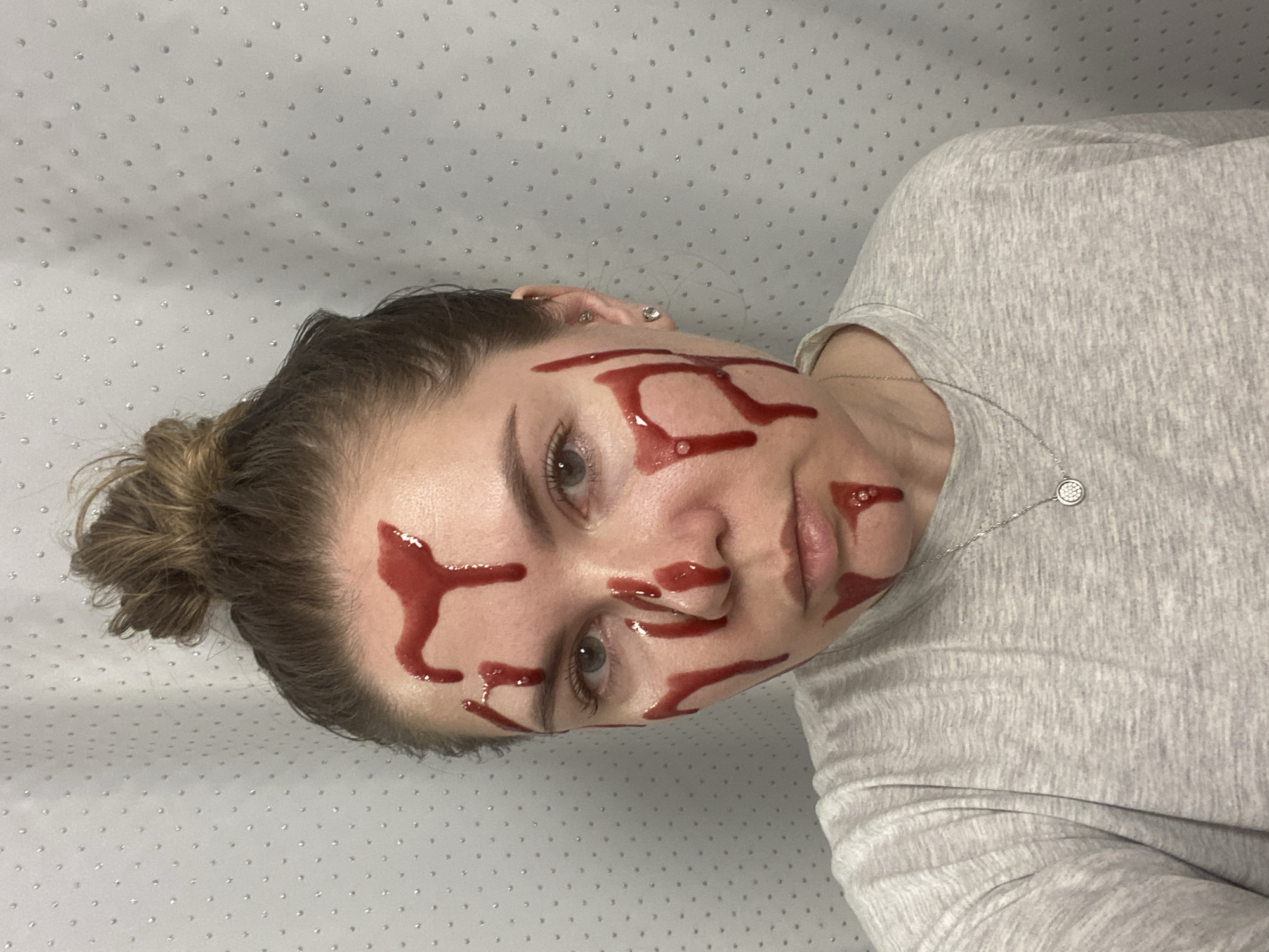 An indoor portrait captures a woman posing with her head tilted to the right, looking directly at the camera. Her face is adorned with numerous streaks and drips of a dark red liquid, resembling artificial blood, which covers her forehead, cheeks, nose, and chin. She has light-colored eyes and dark hair styled in a bun. She is wearing a light grey marled t-shirt and a delicate silver necklace with a circular, patterned pendant. Several small, silver-toned earrings are visible on her right ear, including a stud and a larger circular one. The background is a light, possibly white or very light grey, surface patterned with evenly spaced, small, reflective dots. The overall impression is that of a staged or artistic shot, possibly for Halloween or a similar theme, given the intentional application of the red liquid. No text is visible in the image.