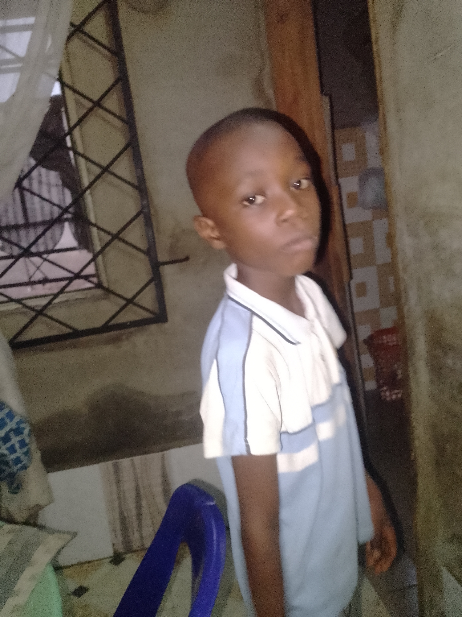 A young, dark-skinned boy stands indoors, facing the camera. He has a closely shaved head and wears a light blue and white collared shirt. Behind him, to his left, is a window with a metal grate. To his right, a partially visible wall with tiled sections and a wooden door frame. Below him, a blue plastic chair. The location is unknown, as no identifying details are present in the image.