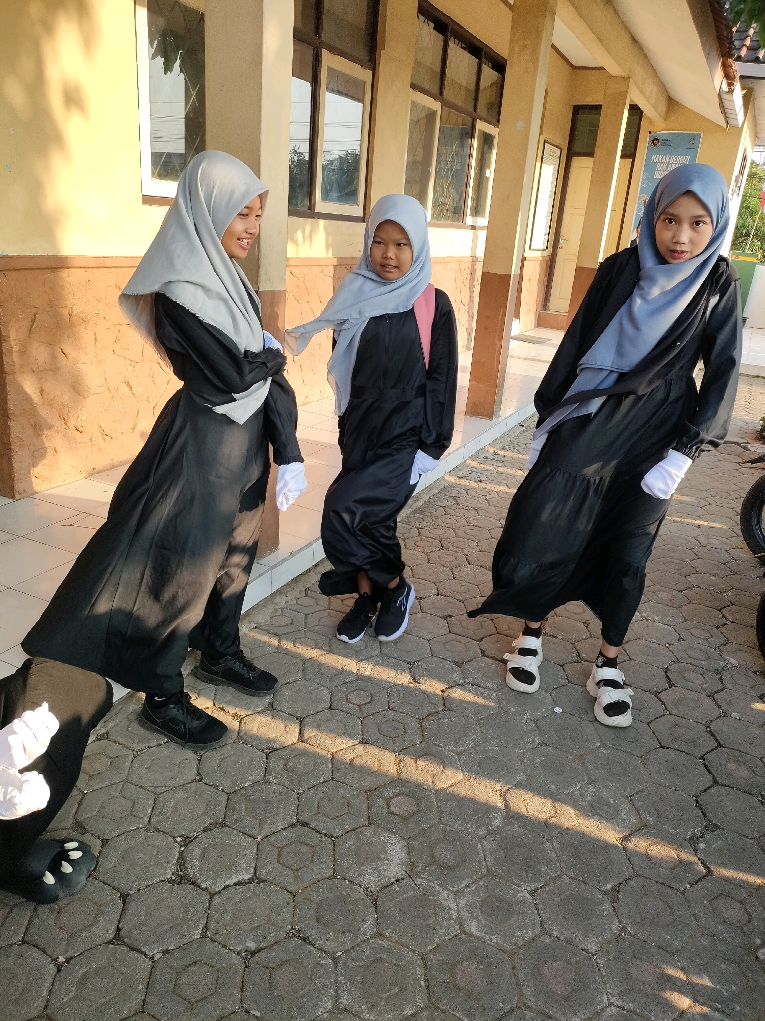 Three young girls in hijabs and long black dresses stand on a paved walkway in front of a building with yellow walls and brown trim. The girl on the left wears a light grey hijab and is smiling. The middle girl, also in a light grey hijab, has a pink backpack and is looking at the camera. The girl on the right, wearing a blue hijab, is looking forward and appears to be posing. All three girls are wearing gloves. The background shows windows, doors, and a sign on the building. The scene appears to be outdoors in a school or community setting.