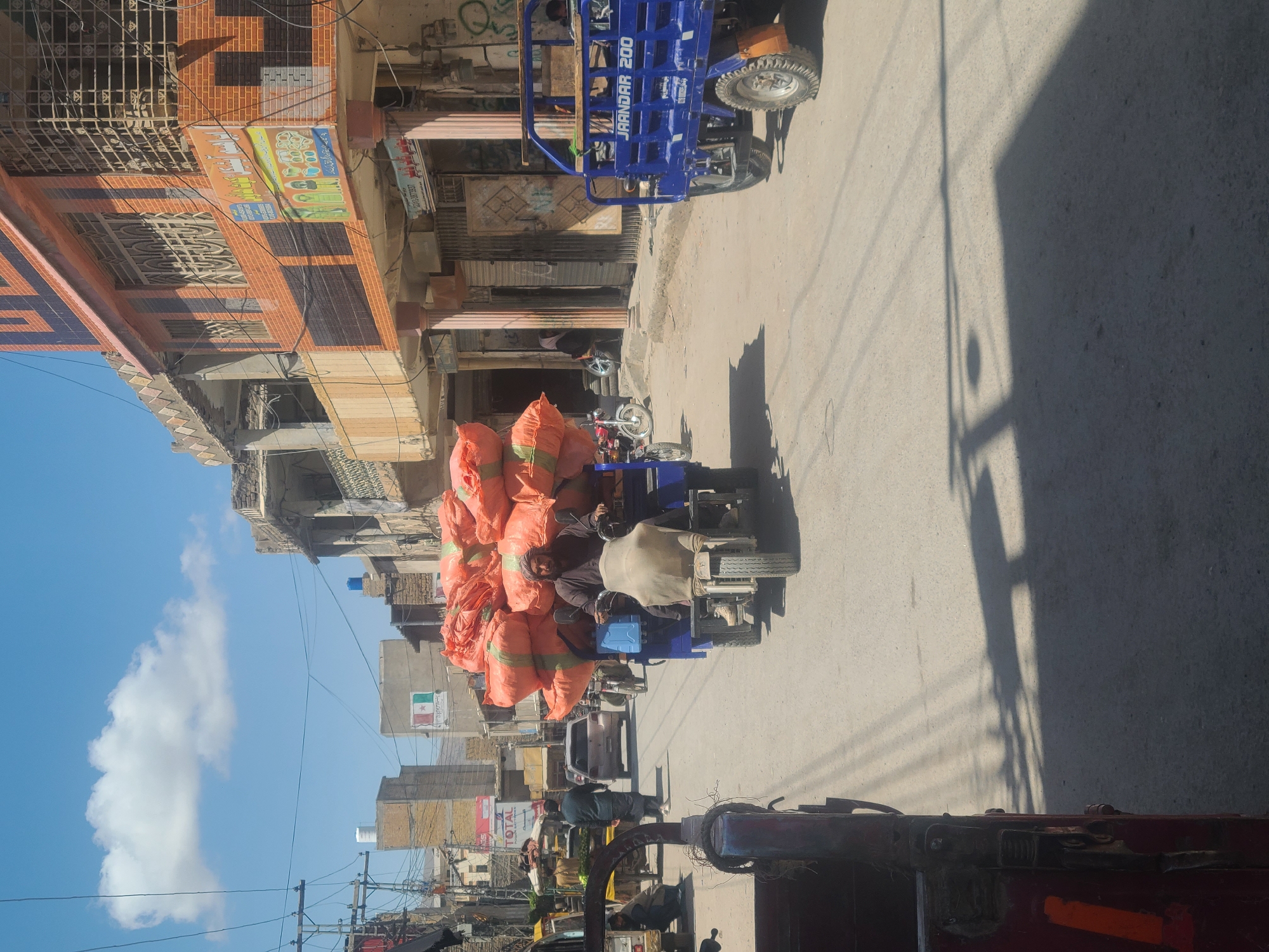 The image depicts a street scene in Quetta City Tehsil, Pakistan. A blue "JAANDAR 200" motorized tricycle is heavily loaded with numerous orange sacks. A man is driving the tricycle, and he is wearing a dark-colored shirt and what appears to be a head covering. The street is paved, and shadows are cast on it. Buildings line the street, with some appearing to be in a state of disrepair. Power lines are visible overhead. Other vehicles and pedestrians are present on the street.