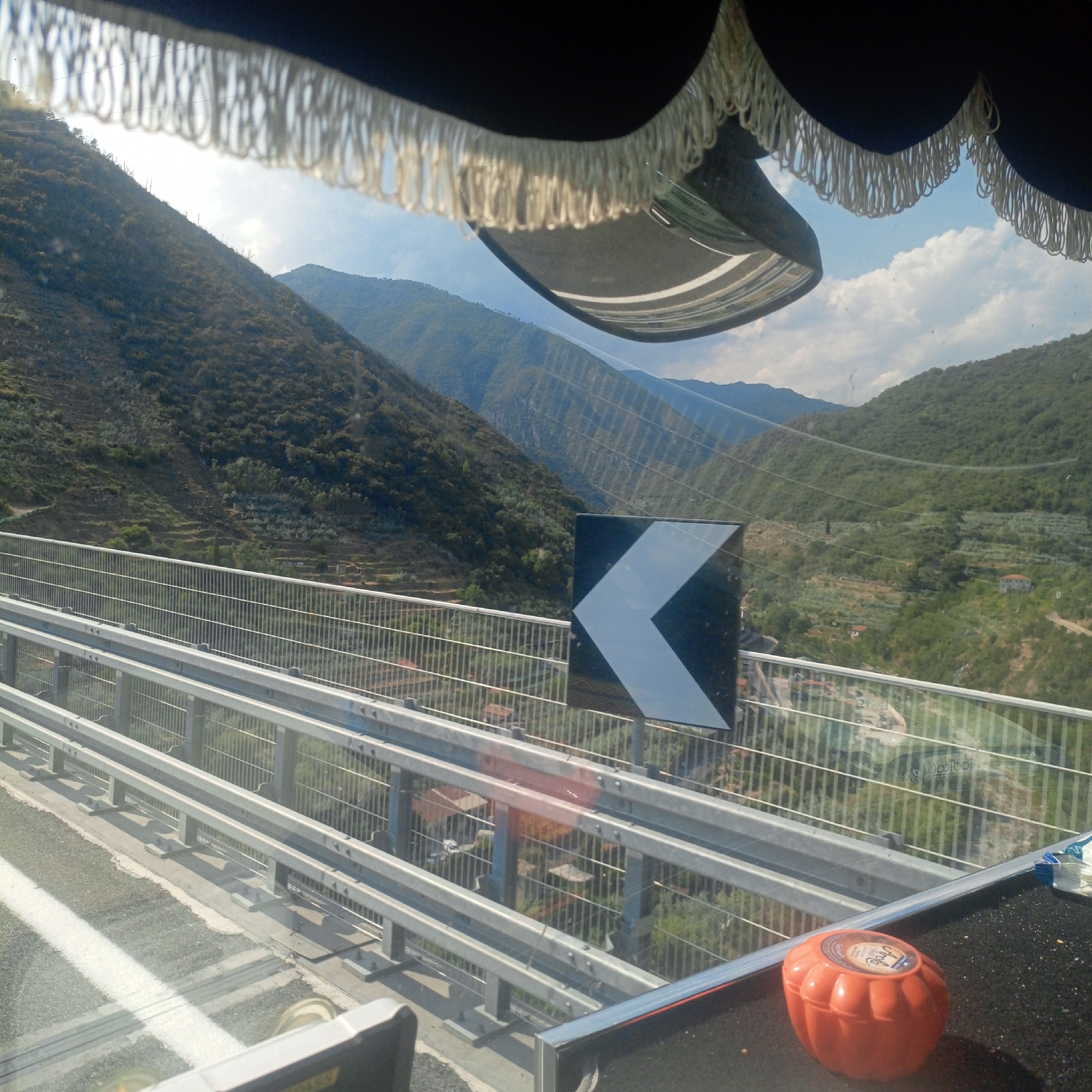 The image appears to be taken from inside a vehicle traveling on a highway overpass. In the foreground, there is a dark-colored interior trim with white fringe hanging down, obscuring the top part of the view. Below this, a shiny surface, possibly a dashboard or interior panel, reflects the sky and the road ahead.
The road itself has white lane markings and a gray asphalt surface. Guardrails made of metal poles and horizontal beams run along the side of the overpass.
Beyond the guardrails, a steep, mountainous landscape is visible. The hills are covered with dense vegetation, appearing as a mosaic of green and brown. Terraced slopes with vegetation are carved into the hillsides, suggesting agricultural use. Several small buildings or houses are scattered throughout the landscape.
A prominent black and white chevron sign, indicating a sharp left turn, is mounted on a pole to the right of the road.
In the far distance, more mountains are visible, fading into a hazy blue. The sky is partly cloudy with bright sunlight.
On the right side of the image, on the surface below the guardrail, a small, orange, pumpkin-shaped object with a label on top is visible.
While the specific location of Taggia, Italy is mentioned, the visual elements themselves do not provide direct confirmation of this location. The landscape is consistent with mountainous regions of Italy.
