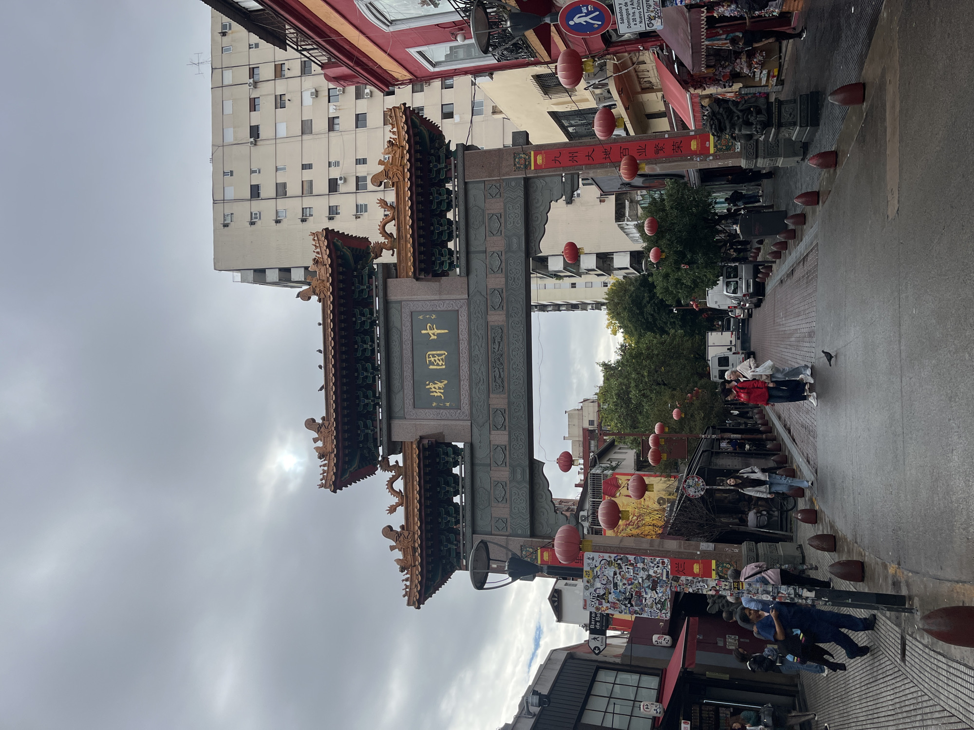 This image depicts a bustling street scene in a Chinatown, specifically in Buenos Aires, Argentina, under an overcast sky during daytime. The focal point is a grand, traditional Chinese archway (paifang) spanning the street, adorned with intricate golden characters and dragon sculptures on its tiered roofs. The characters "城國中" (Chinatown) are prominently displayed on the archway. Red banners hanging from the archway feature the text "九州大地百业繁荣," which translates to "The land of Kyushu prospers in all industries."
Numerous red Chinese lanterns are strung across the street and hang from poles, adding to the festive ambiance. Pedestrians can be seen walking along the street and sidewalks. On the right side, the sidewalk is lined with red, dome-shaped bollards. Various buildings, including storefronts with awnings and a tall, plain apartment building, line the street. White vans are visible parked on the left side.
The sky is largely overcast, though a bright patch of diffused sunlight suggests the sun is trying to break through. Visible text on a street sign includes "Sábados y Domingos de a 20 hs. y Añ Nuevo Chin lo Ingre," likely referring to event hours or admission information for the Chinese New Year. Another sticker-covered pole displays "Barrar SKATE de Bel." The overall scene captures the vibrant daily life of a cultural district.
