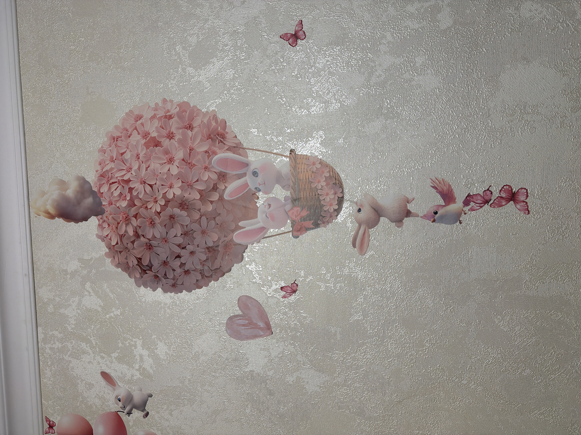 This image depicts a wall decoration featuring a whimsical scene with bunnies and a hot air balloon. The dominant element is a large, spherical hot air balloon constructed from pink flowers, with a fluffy cloud-like top. Two bunnies are seated in a woven basket attached to the balloon. Below the balloon, another bunny is depicted in mid-air, followed by a small pink bird perched on a branch, and two pink butterflies. To the left of the balloon, several pink balloons are being held by a bunny, and a pink heart floats nearby. The background is a textured wallpaper in a light, neutral tone. The scene is entirely decorative and appears to be a wall decal or mural. The location is identified as Unknown, Unknown.