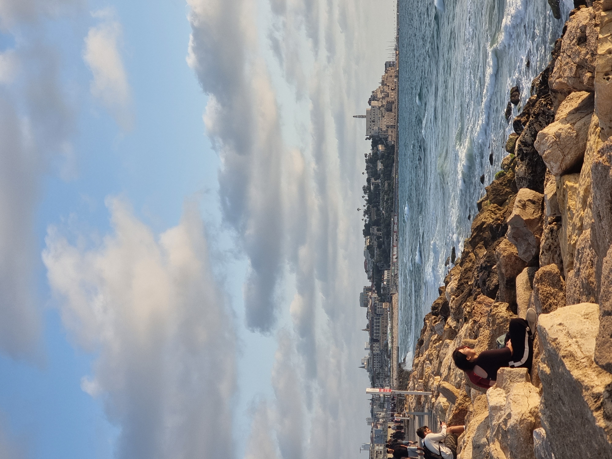 The image presents a scenic coastal view, likely Tel-Aviv, Israel, captured during the late afternoon or early evening. The photograph is composed with a rocky foreground, the sea and a city skyline in the middle and background, and a partially cloudy sky overhead.
In the lower right foreground, a young woman with dark hair, dressed in a black top and dark pants with white side stripes, is seated on large, light-colored rocks. She appears relaxed, looking out towards the sea and the distant urban landscape. To her left, on a paved promenade, several other people are visible walking or standing, enjoying the coastal atmosphere.
The mid-ground is dominated by the blue-green sea, featuring moderate waves actively breaking against the rocky shoreline. The city skyline stretches across the horizon in the background, displaying numerous buildings of varying heights and architectural styles, including a prominent taller structure, possibly a minaret or historical tower, rising above the cityscape.
The sky, occupying the upper portion of the frame, is partly cloudy, with patches of clear blue visible between large, soft grey and white clouds that extend across the breadth of the image. The lighting is soft and warm, characteristic of a golden hour, illuminating the scene with a pleasant, tranquil ambiance. A red and white sign on the promenade displays the partial text "Cig". The overall scene depicts a blend of natural coastal beauty and urban development, suggesting a popular recreational area.