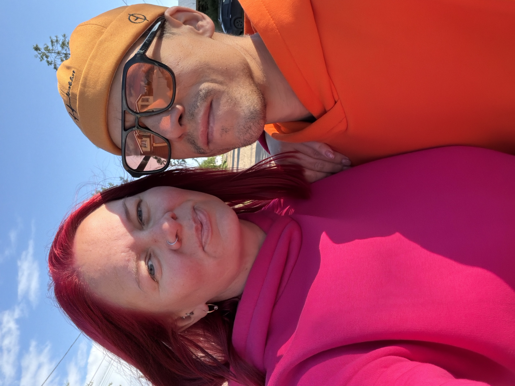 This is an outdoor selfie featuring two individuals, a man and a woman, captured on a sunny day. The image is rotated 90 degrees clockwise, showing the subjects from a low angle against a bright blue sky with scattered white clouds.
The man, positioned in the upper left of the rotated image, wears an orange beanie with a black circular symbol (possibly an anarchy symbol) and the word "BEANZ" embroidered on it. He also sports dark-framed sunglasses with light pinkish lenses, which reflect a building with a red roof. He has light facial stubble and a relaxed expression, wearing a vibrant orange hooded sweatshirt.
The woman, in the foreground on the lower right, has bright red hair that frames her face. She wears a silver nose ring and several earrings, including one shaped like a safety pin. She also wears a vivid pink hooded sweatshirt. Her hand, with light-colored nail polish, is visible, resting on the man's arm or shoulder. She looks directly at the camera with a neutral to slightly smiling expression.
The bright sunlight casts noticeable shadows, indicating a clear, sunny day, likely during the late morning or afternoon. The background details are sparse due to the close-up, low-angle shot, but hints of green foliage and what appears to be a residential street or trees can be seen. The scene conveys a casual and pleasant moment. No specific landmarks for Chaikovsky, Russia, are visible, but the setting is consistent with an urban or suburban outdoor environment.