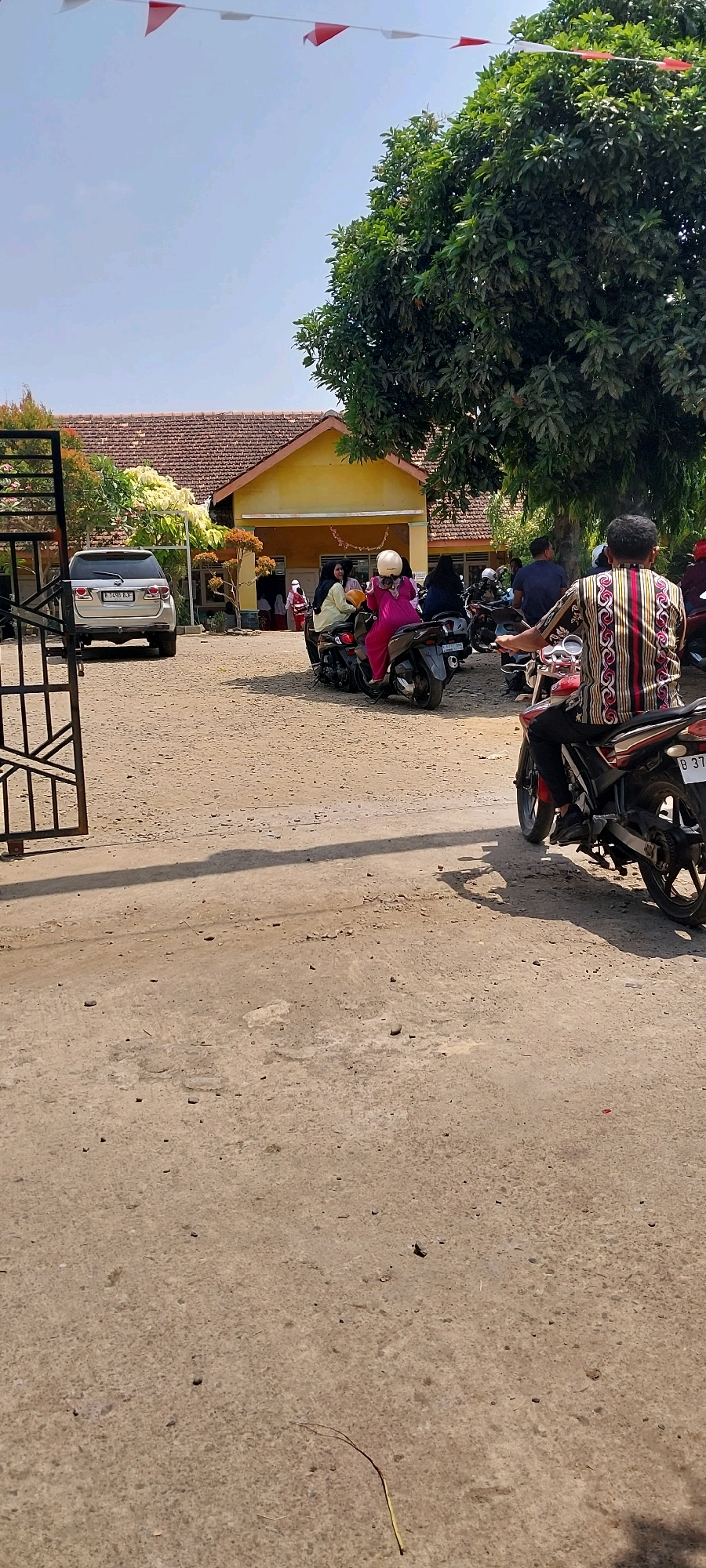 The image captures a sunny day scene outside a yellow, single-story building with a tiled roof in Keling, Indonesia. Red and white triangular flags are strung across the top of the frame, suggesting a festive occasion. In front of the building, several people are gathered. A silver SUV is parked on the left. A black metal gate is visible on the far left, with its shadow cast on the sandy ground. A group of people are on motorcycles, with some being driven and others as passengers. One individual in the foreground is on a motorcycle, wearing a patterned shirt and black pants. Another person on a motorcycle to their left wears a pink dress and a white helmet. The ground is unpaved and appears dusty. Lush green trees are present to the right of the building and in the background, contributing to the tropical feel of the location. The overall atmosphere appears to be one of community gathering or event at the building.