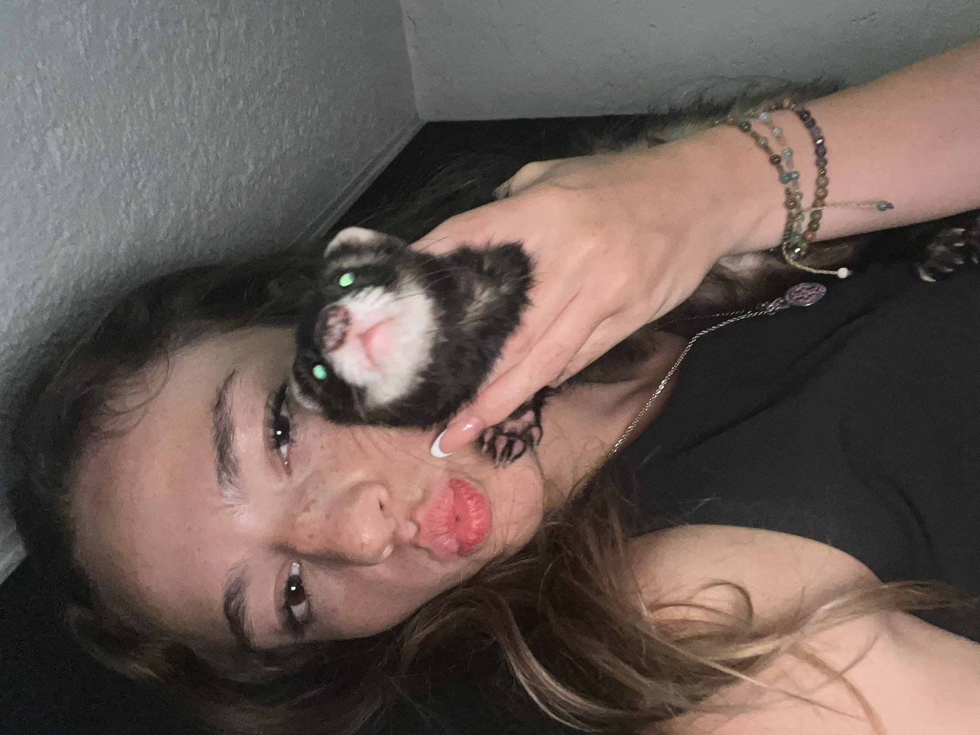A woman is lying down, cradling a ferret in her hand. The ferret's head is near the woman's face, with its pink nose and green eyes visible. The woman has light skin, freckles, and brown hair. She is wearing a black top and a beaded bracelet. The background is a plain, light-colored wall. The location is unknown.