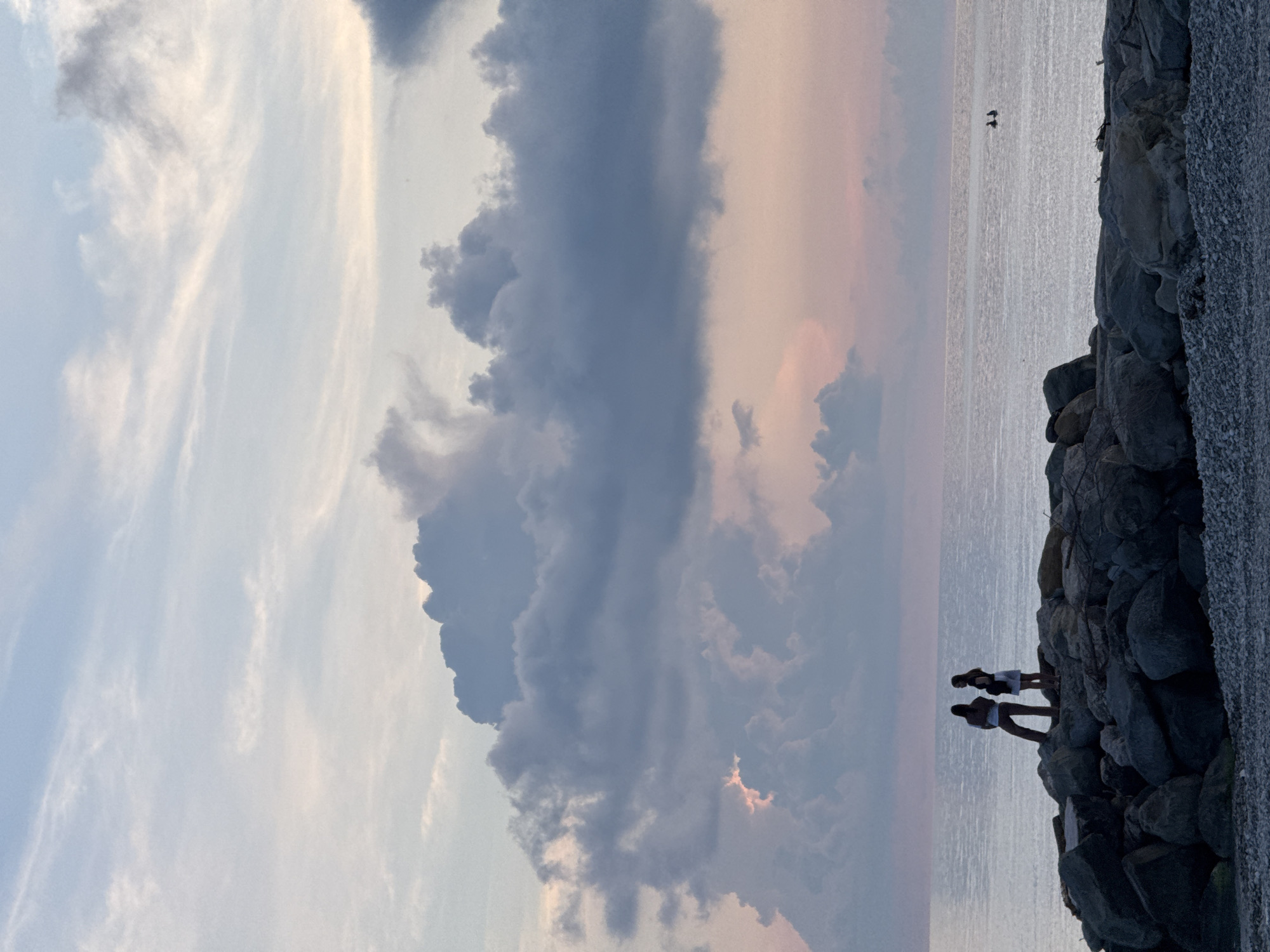 The image depicts a scenic view of the Black Sea coastline near Nebug, Russia, during what appears to be either sunrise or sunset. The sky is a canvas of soft blues and pinks, with dramatic, heavy clouds dominating the upper portion of the frame. Wispy cirrus clouds are scattered across the lighter parts of the sky.
In the foreground, a rocky breakwater is visible, composed of large, irregularly shaped stones. On top of this breakwater, two figures are silhouetted against the sea and sky. They appear to be standing and facing away from the camera, looking out towards the water. One figure is taller and appears to be wearing pants and a shirt, while the other is shorter and seems to be wearing a dress or skirt. Their postures suggest a moment of quiet contemplation or observation.
The sea is calm and reflects the muted colors of the sky. In the distance, on the water, two small, dark shapes are discernible, which could potentially be kayakers or small boats. The overall atmosphere is serene and picturesque, characteristic of a coastal landscape at the transitional hours of the day. The pebbled shore is visible at the very bottom of the image.