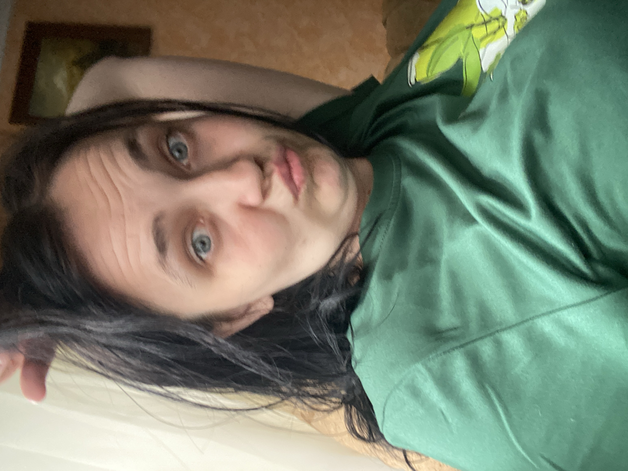 A woman with dark hair is leaning back, her left arm behind her head, and her right arm is not visible. She is wearing a green t-shirt with a white and green graphic on the right side of the chest, partially obscured by her arm. Her eyes are wide and blue, and her lips are pursed. The background includes a cream-colored curtain to the left and a brown wall with a framed picture to the right. The scene appears to be indoors, possibly in a living room or bedroom.