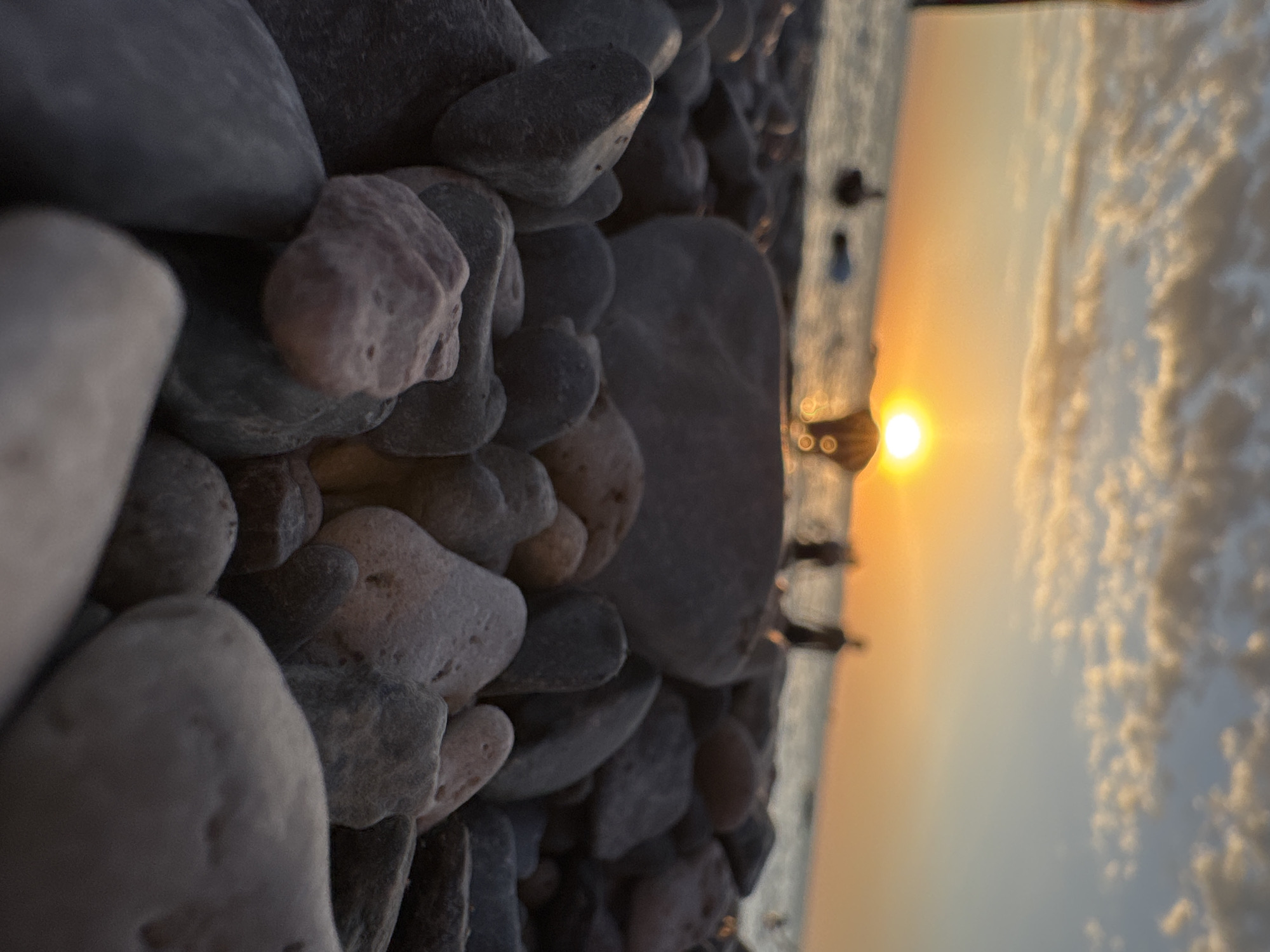 The image is a low-angle, close-up shot of a pebble beach at sunset. The foreground is dominated by smooth, grey and pinkish rocks, some of which are in sharp focus while others are blurred. Beyond the rocks, the calm sea is visible, reflecting the warm colors of the sky. In the distance, silhouettes of people can be seen standing in or near the water, and on the beach. The sun is setting on the horizon, casting a golden glow and creating a halo effect. Wispy clouds are scattered across the sky, tinged with orange and yellow. The scene evokes a sense of tranquility and the end of a day at the beach, with the location being Zharbulak, Kazakhstan.