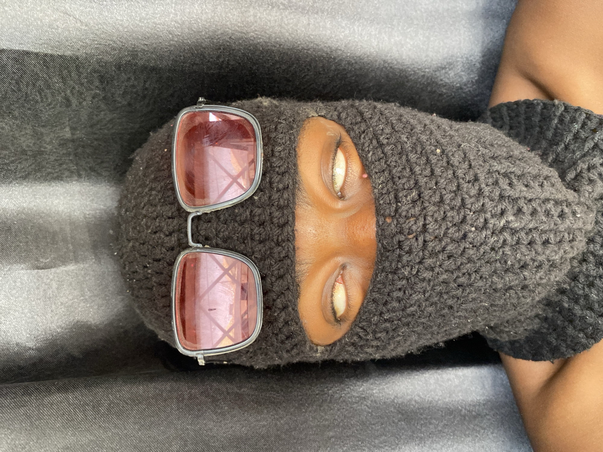 A person wearing a dark gray knitted balaclava with narrow slits for the eyes is visible. The person's skin tone is brown. Rectangular sunglasses with rose-colored lenses and a silver frame sit askew on the balaclava. The background appears to be a gray fabric. The context regarding Barakin Akawo, Nigeria, is not provided.