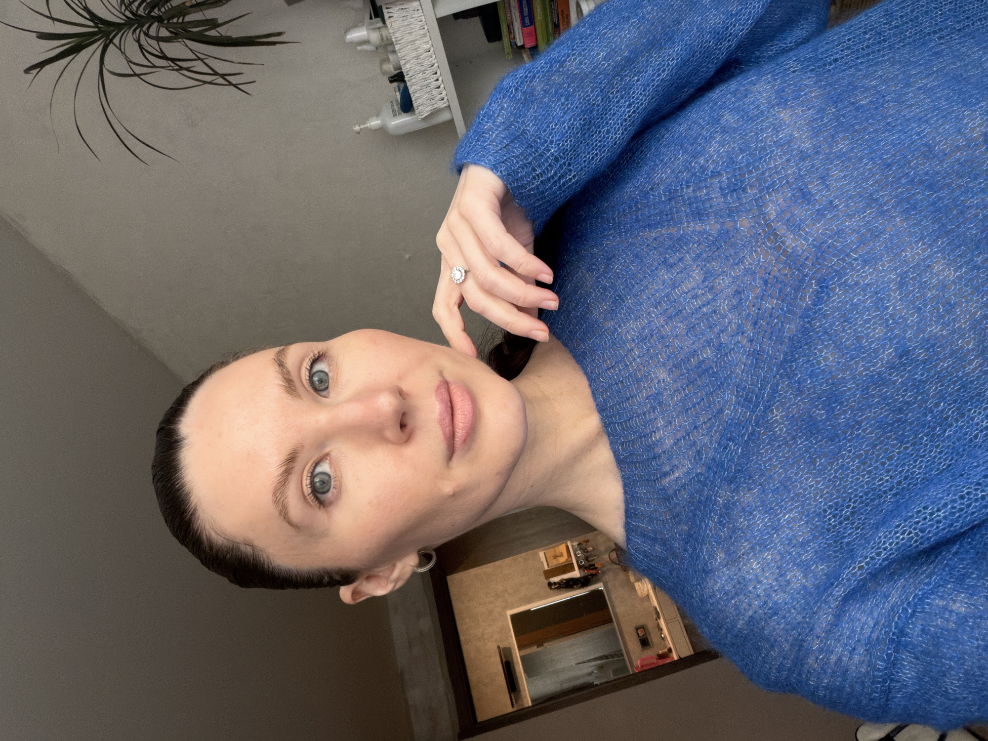 A woman is posing for a selfie indoors. She has fair skin, brown hair pulled back, and blue eyes. She is wearing a bright blue sweater and a ring is visible on her left hand. In the background, there is a glimpse of a doorway leading to another room. Shelves with various items are visible in the upper left. The location appears to be a private residence, not identifiable to Perm, Russia.