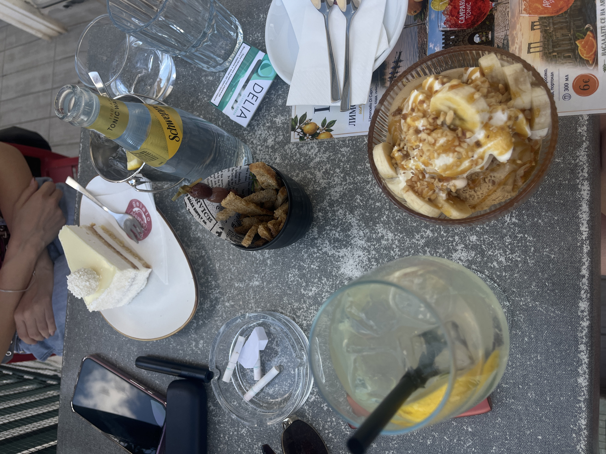 A high-angle outdoor shot shows a table setting with food and drinks. In the foreground, a glass of lemonade with ice and a lemon slice sits on the table, accompanied by a dessert bowl filled with ice cream, bananas, and nuts. To the right, a menu shows a picture of an orange drink. Further back, a slice of white cake with coconut shavings is on a plate. A bottle of Schweppes tonic water rests in an ice bucket. An ashtray contains cigarette butts. A smartphone and a pack of IQOS cigarettes are also present. In the background, two people are partially visible, sitting at the table. The setting appears to be a cafe or restaurant.
