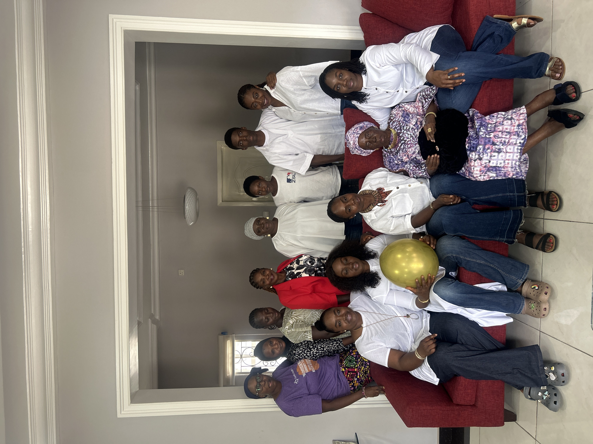 A group of 12 individuals, including men and women of various ages, are posing for a photograph in what appears to be a living room. The group is arranged in two rows on a red couch and in front of it. The individuals are dressed in casual and formal attire, with some wearing traditional Nigerian clothing. One person in the front row is holding a golden balloon. The room has light grey walls and a white trim around a doorway. The lighting appears to be natural, coming from an unseen source. The image quality is clear, allowing for detailed observation of the people and their surroundings. The scene is a family gathering or celebration.