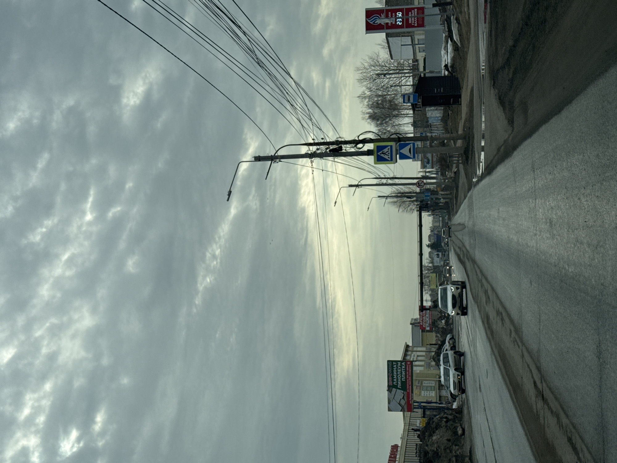 The image depicts a street in Chaikovsky, Russia. The view is along a road with several cars visible. The sky is overcast with heavy cloud cover. Power lines stretch across the sky. Sidewalks line the road. Buildings and billboards are present on the side of the road. Roadside debris and construction or repair materials can be seen in places. A pedestrian crossing sign is visible on a pole.