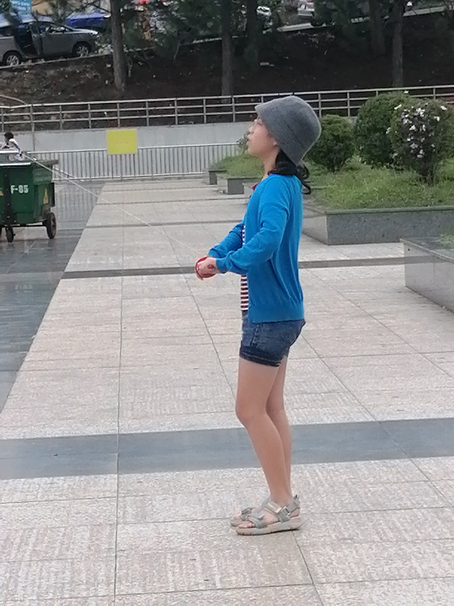 A young girl wearing a grey knitted hat, blue cardigan, and denim shorts stands on a paved pathway. She is holding a red object in her hands, which appear to be positioned in front of her body. To the left of the girl is a green refuse bin with "F-85" visible on its side. In the background, there is a railing, some trees, and bushes. The scene appears to be an outdoor urban environment, possibly a park or a public walkway.