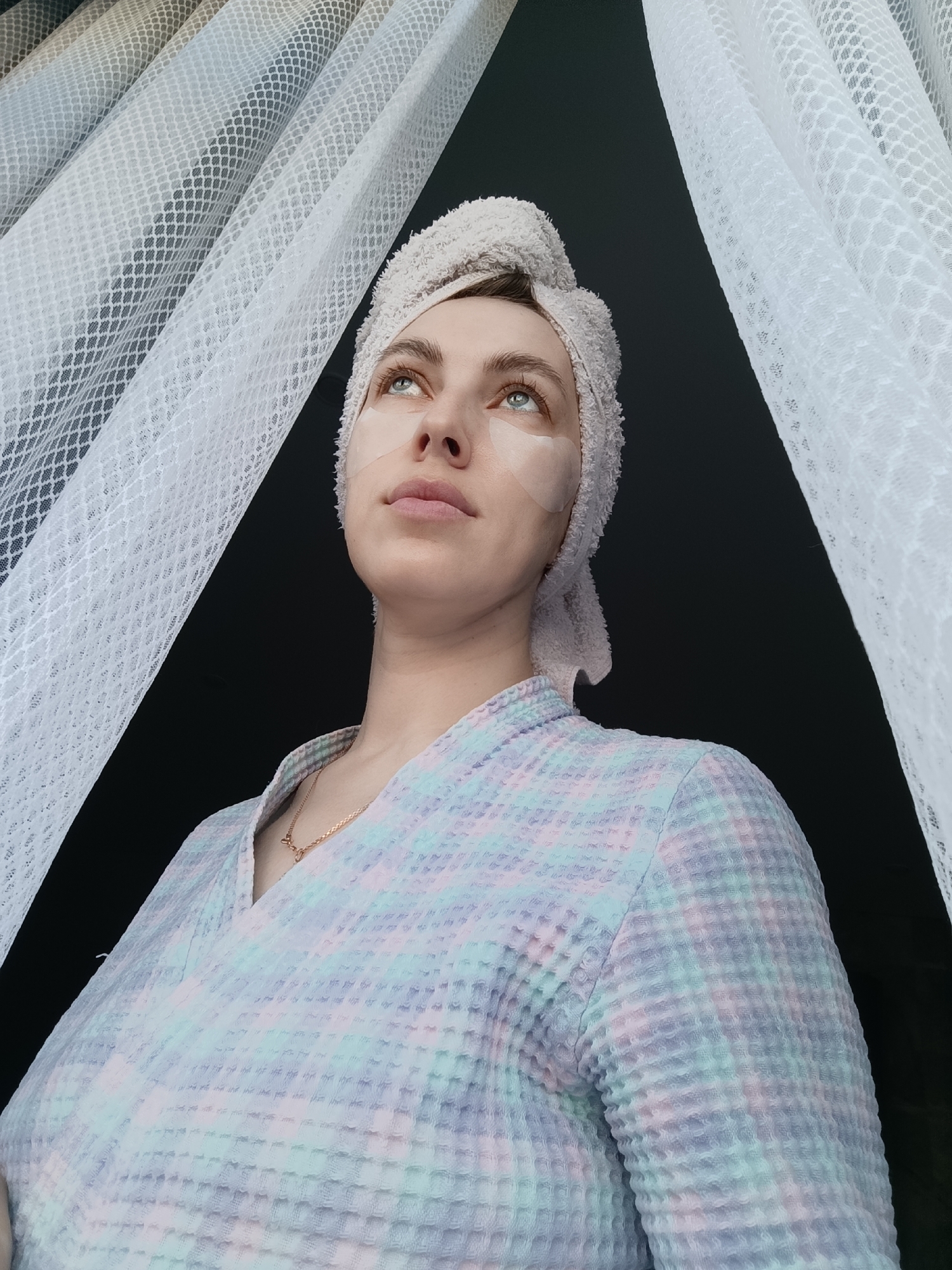 A low-angle medium shot captures a fair-skinned woman with a towel-wrapped head and under-eye masks. She is wearing a light-colored, waffle-patterned robe and a gold necklace. She gazes upwards and slightly to the left. A white, net-like curtain is visible in the upper left of the frame, contrasting against a dark background. This scene appears to have been taken indoors, potentially in Chaikovsky, Russia.