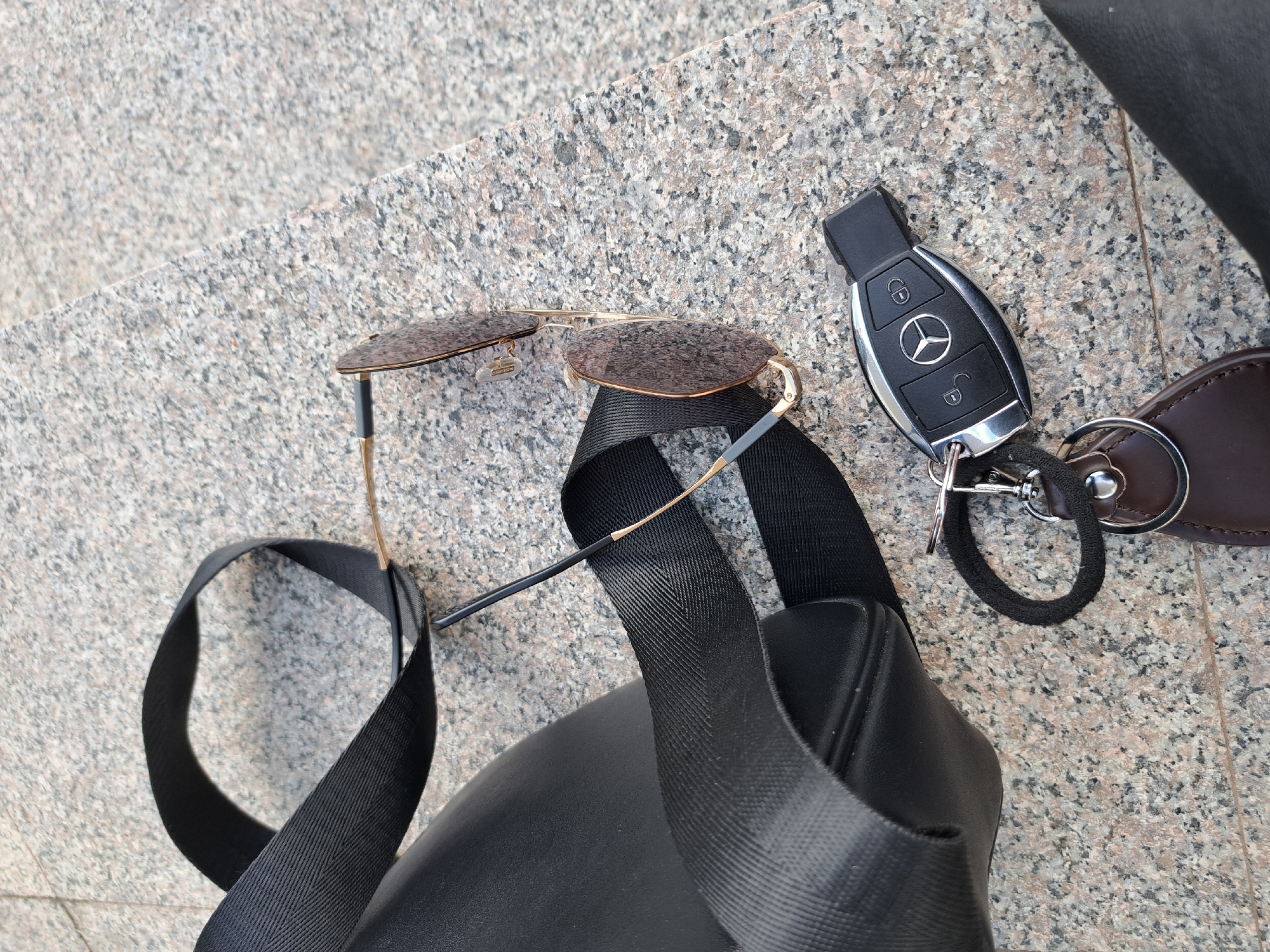 An overhead shot captures a collection of personal items resting on a light grey, speckled granite surface, likely an outdoor pavement or step in Negovan, Bulgaria. The main subjects are a pair of aviator-style sunglasses, a Mercedes-Benz car key, and a black bag with its strap.
The sunglasses, with gold-toned frames and dark, possibly brown-tinted lenses, are positioned centrally, partially obscuring a black woven strap that appears to belong to the bag. A small tag is visible on the left lens hinge of the sunglasses, bearing the text "BLANCHE". The Mercedes-Benz key fob is situated to the right of the sunglasses, displaying the iconic three-pointed star logo and symbols for locking and unlocking a vehicle. It is attached to a keychain that includes a dark brown leather tab, a black coiled cord, and a silver-colored metal ring and clasp. The black bag, made of what appears to be leather or a similar material, is partially visible in the lower right and left corners of the frame, with its textured strap looping across the granite.
The lighting suggests it is daytime, casting no harsh shadows. There are no people visible, and no specific activity is taking place other than the items being laid out.