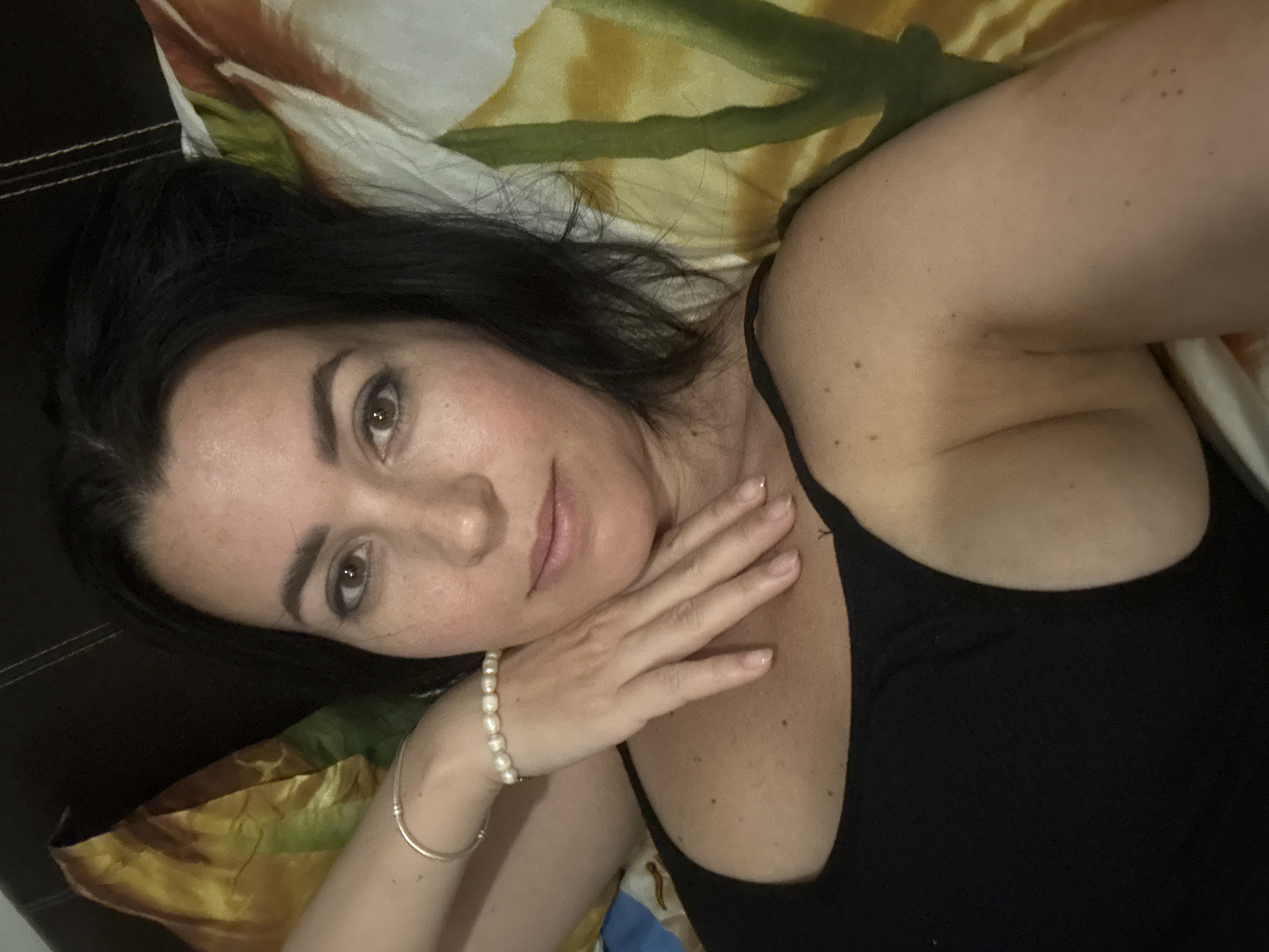 A woman with dark hair lies on a patterned fabric surface, possibly a bed or couch, with her head tilted slightly towards the camera. She is wearing a black tank top and a pearl bracelet on her left wrist. Her left hand is positioned near her face. The fabric beneath her features a pattern with yellow, green, and white colors. The image does not provide any information that would confirm the location in Pattaya City, Thailand.