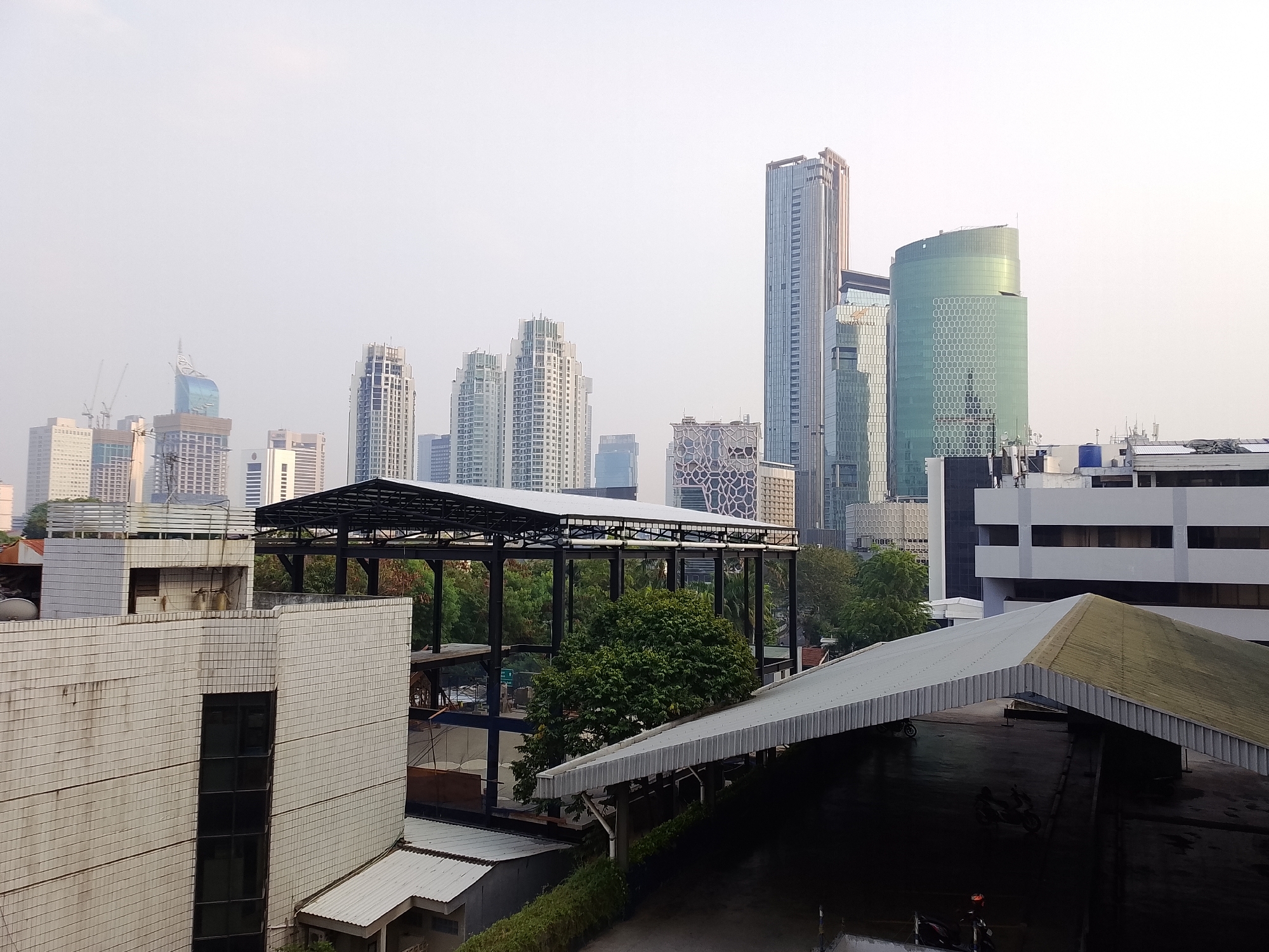 This is a wide-angle, slightly elevated, eye-level shot of a cityscape in South Jakarta, Indonesia, likely taken during the daytime with overcast skies contributing to a muted color palette. In the foreground, a white tiled building with a dark rectangular window is visible on the left. To the right, a large, sloping, gray corrugated metal roof extends into the frame, covering an area that appears to be a covered parking lot or outdoor space, with two motorcycles parked beneath it. Behind these structures, a black metal-framed open-air building or canopy is under construction or renovation, with trees growing through it and in the background.
Further back, the mid-ground and background are dominated by a dense collection of modern skyscrapers of varying heights and architectural styles. Notable buildings include a tall, slender skyscraper with a distinctive pointed, blue-tinted top, and several other high-rise buildings with glass facades and repeating geometric patterns. The overall impression is one of urban development and a bustling city environment. The lighting is soft and diffused, suggesting the photo was taken at dawn, dusk, or on a heavily overcast day. There are no visible people in the frame.