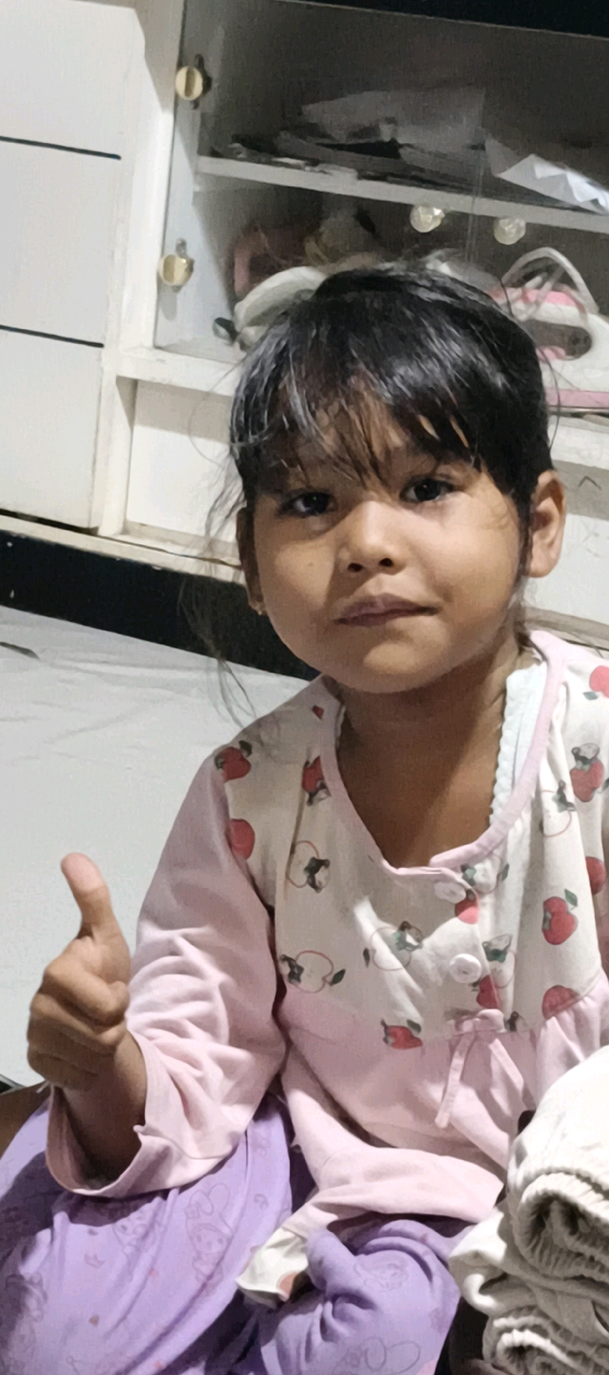 A young girl is sitting in front of a white cabinet with open shelves in a dimly lit room. The cabinet contains various items, including what appear to be shoes, bags, and other miscellaneous objects. The girl is wearing a pink long-sleeved shirt with a fruit pattern and purple pants. She is giving a thumbs-up gesture with her left hand. Her hair is dark and styled with bangs. The background shows a white wall or furniture piece to the left of the cabinet. The overall environment suggests an indoor setting, possibly a home. The location is stated to be Muaro Jambi, Indonesia.
