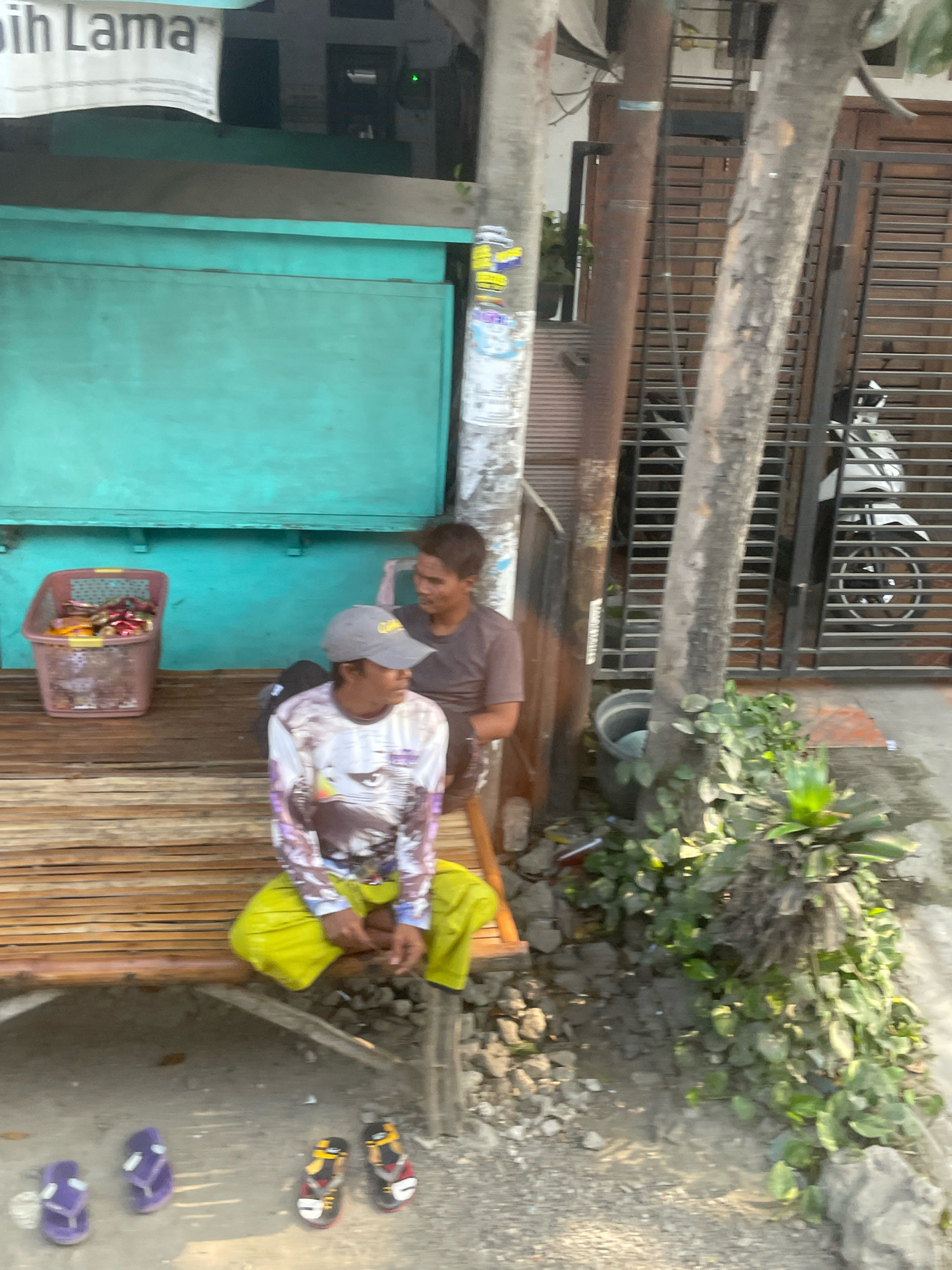 This image captures a street scene in Kramat, Indonesia. Two individuals are seated on a wooden bench. One person, wearing a grey baseball cap and a long-sleeved shirt with an eagle graphic and yellow pants, is facing to the right. The other individual, wearing a dark t-shirt and sitting closer to the right edge of the bench, is looking towards the left. In front of the bench, there are three pairs of flip-flops: two purple pairs on the left and one multi-colored pair on the right. To the left of the bench, a plastic basket filled with items, possibly food or goods, is placed on the ground. Behind the bench, a turquoise-colored structure with a dark horizontal beam is visible, and a white banner with the text "ih Lama" is attached to the top of this structure. To the right, a tree trunk stands prominently next to a metal gate with horizontal bars. A white motorcycle is partially visible behind the gate. The ground is a mixture of dirt, gravel, and some vegetation growing along the base of the tree and the gate. The overall scene suggests a casual, possibly commercial, outdoor setting.