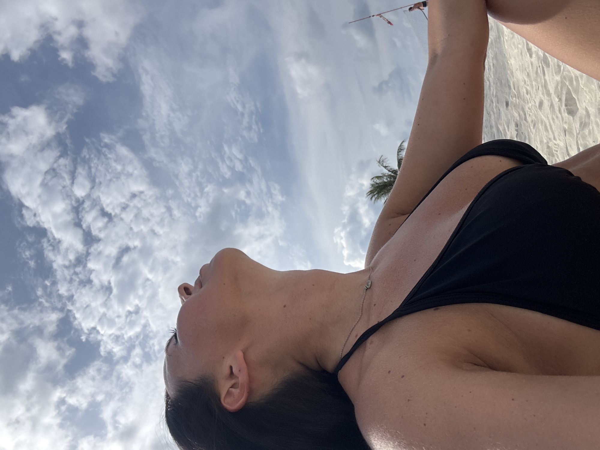 A low-angle shot reveals a woman with dark hair, wearing a black bikini, lying on a beach. A thin silver necklace rests on her chest. Above, a sky filled with white clouds is visible, along with a palm tree and a distant object resembling a volleyball net. The sandy beach can be seen to the right of the woman. The scene appears to be set in Baan Chaloklum, Thailand.