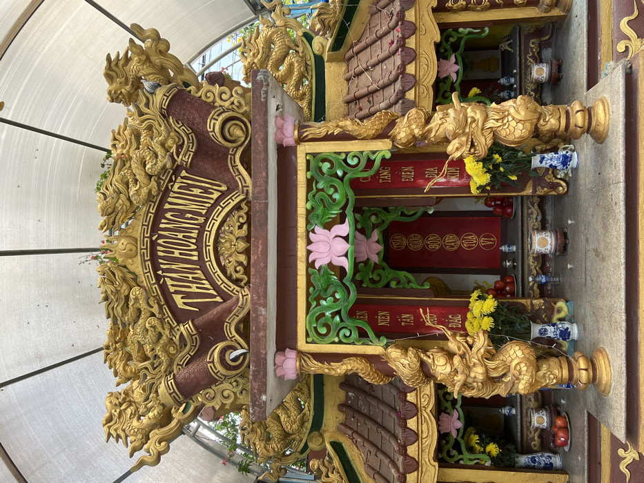 This image depicts a highly ornate, multi-tiered altar structure identified by the prominent gold Vietnamese inscription "THẦN HOÀNG MIẾU" on a red plaque at its apex, located in Đăk Mar, Vietnam. The structure is predominantly gold, red, and brown, featuring extensive sculptural details.
The uppermost section is adorned with multiple golden dragon figures intricately carved and facing outward. Below this, a reddish-brown tiled roof section with textured details is visible. The central frontal area of the altar is an open recess framed by gold and green decorative elements. A large pink lotus motif is centered above the inner opening, which features red panels with vertical gold Vietnamese script.
Two golden dragon sculptures serve as pillars flanking the main altar entrance, resting on the lower platform. On the altar platform itself, various items are arranged symmetrically: several vases containing yellow flowers (chrysanthemums), bowls of red fruits (appearing to be apples or pomegranates), and multiple smaller ceramic or porcelain vessels, some with blue and white patterns, potentially functioning as incense burners or offering bowls. Small, unlit candles are also present on the platform.
The entire altar is situated beneath a translucent, arched corrugated roof structure supported by metal framework, providing shelter. Green foliage is visible in the background, suggesting an outdoor or semi-enclosed garden setting. No individuals or active interactions are visible within the frame. The materials show slight signs of weathering and environmental exposure, such as minor discoloration on the roof tiles.