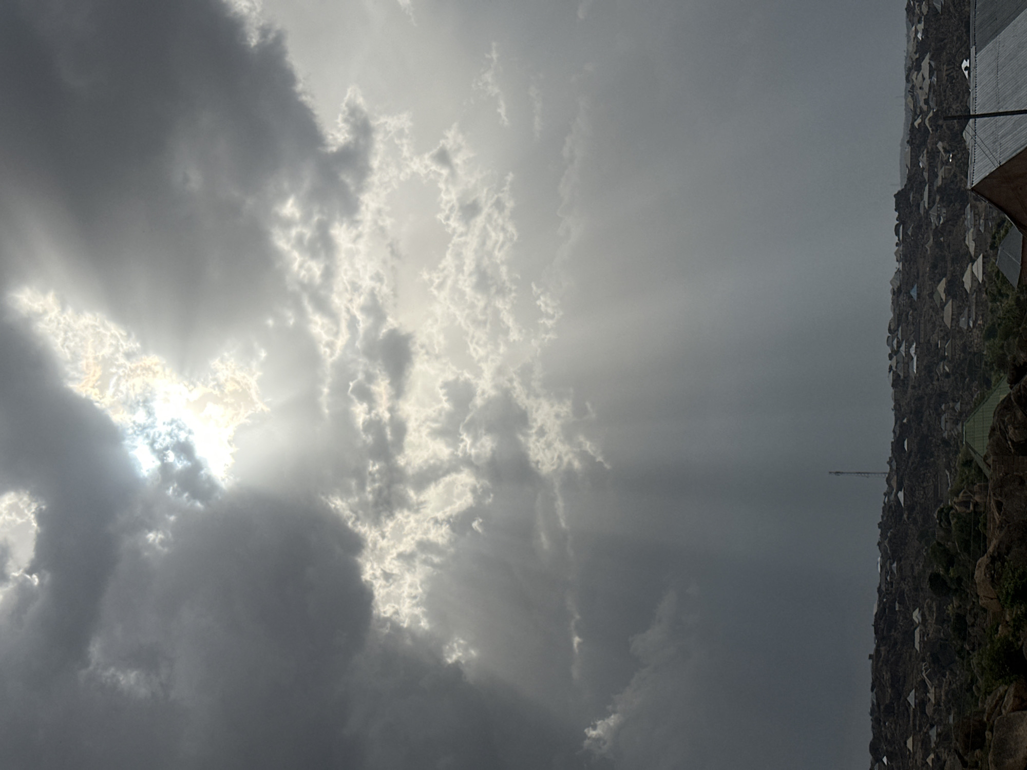 The image shows a cloudy sky with the sun shining through the clouds. The sun's rays are visible and there is a dark land with several buildings in the background. A communications tower rises above the buildings, it is set against the city of Jos, Nigeria.