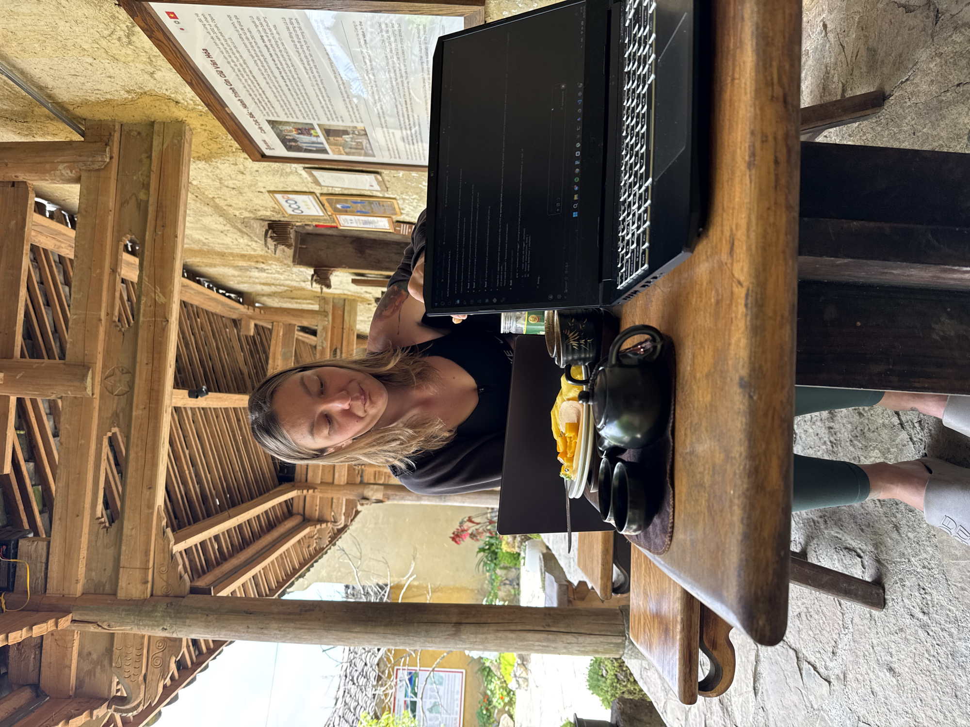 A woman is seated at a wooden table with a laptop in front of her, possibly working or browsing the internet. A tea set, including a teapot and cups, is visible on the table next to the laptop. The setting appears to be an open-air or partially enclosed space, with wooden beams and traditional construction elements visible in the background, suggesting a rural or cultural environment. The image contains text that indicates the location is within the city of Lũng Cú Commune, Vietnam. There is also what appears to be a sign or informational plaque on the wall behind the woman, containing text and small pictures. The floor appears to be stone.