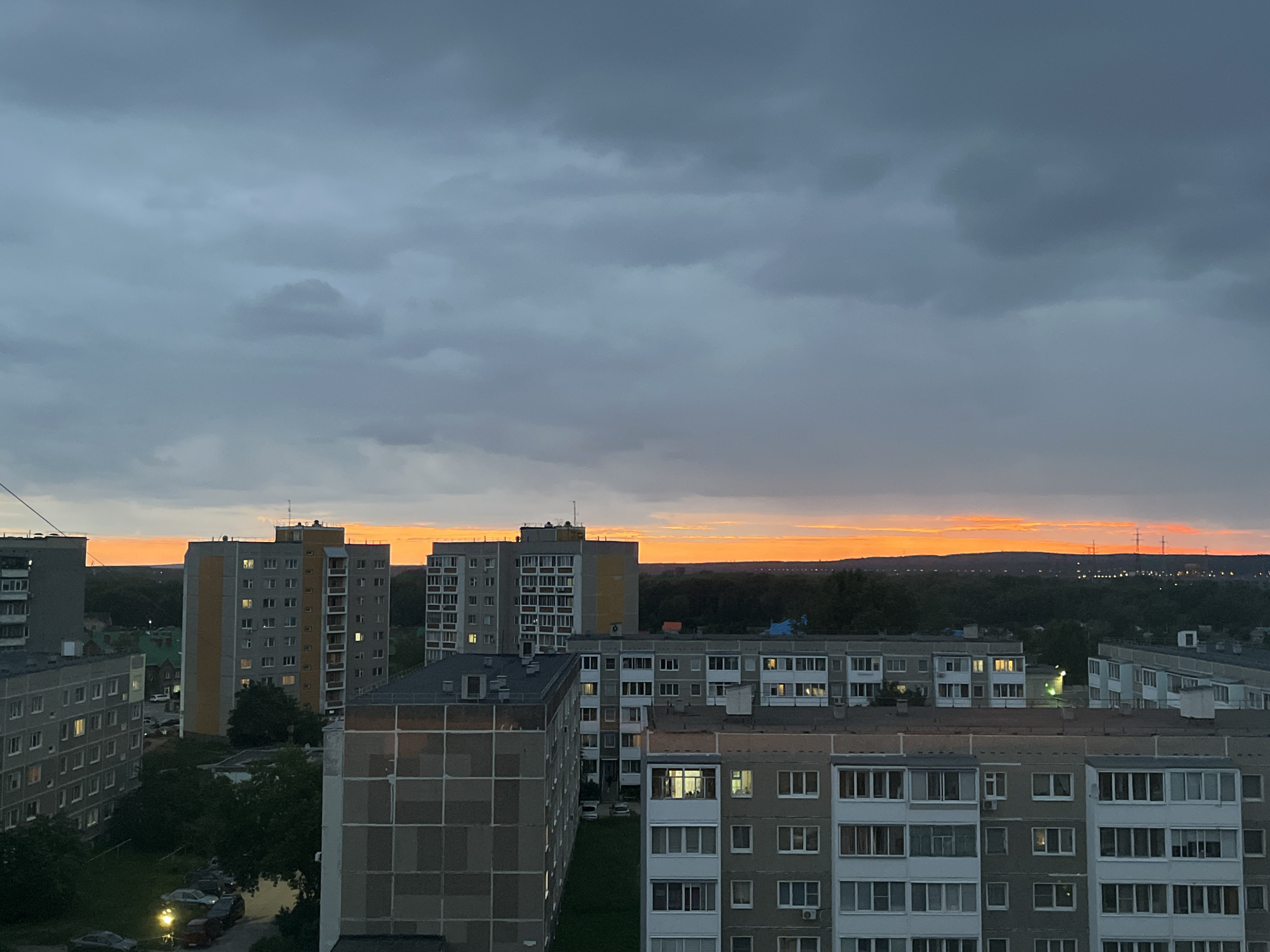 An aerial view shows a dense urban environment at dusk, with a dramatic sunset painting the sky with vibrant orange and yellow hues. In the foreground, several multi-story residential buildings, typical of Soviet-era architecture, are densely packed. These buildings are constructed from concrete and have uniform window patterns. Some windows are illuminated, suggesting occupants are home. Trees and greenery are visible between and around the buildings, indicating a degree of urban planning with natural spaces. Beyond the residential blocks, a darker line of trees forms a natural barrier, with the bright band of the sunset visible above it. In the far distance, a silhouetted treeline is punctuated by the faint glow of distant city lights and what appear to be power lines or transmission towers. The sky is dominated by heavy, dark grey clouds that obscure most of the sky, making the vibrant sunset colors particularly striking. The overall impression is one of a utilitarian, lived-in urban landscape under a dramatic twilight sky.