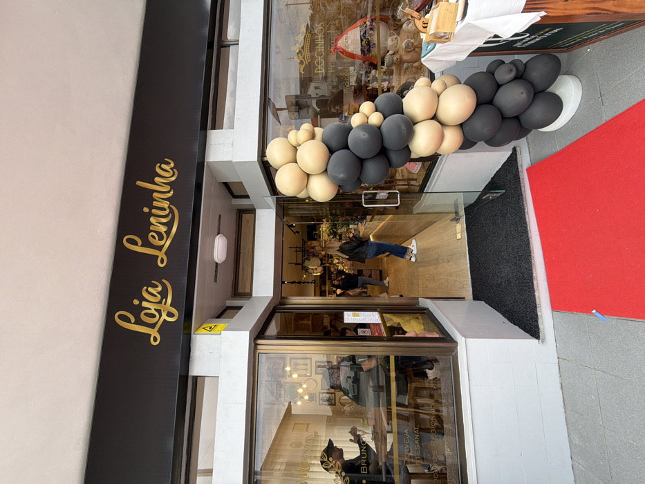 The image depicts the exterior and partial interior of a commercial storefront identified as "Loja Leninha" in Felgueiras, Portugal.
The store's facade features a black sign with "Loja Leninha" inscribed in gold script. Below the sign, a central glass door serves as the entrance, flanked by large glass display windows. A red carpet extends from the threshold onto the gray concrete pavement outside, indicative of a special event or opening. A large, organic-style balloon garland, composed of beige and black balloons of various sizes, is positioned vertically to the right of the entrance, further supporting the event context.
Through the left display window, two individuals are visible seated at a light-colored wooden table. One individual, wearing a dark cap, is seen from behind. Text adhered to the window glass advertises "BRUNCH," "CERVEJA ARTESANAL," and "PRODUTOS REGIONAIS."
Inside the store, visible through the open door and right window, several individuals are present. One woman, wearing a black jacket and blue jeans, stands near a counter area, facing left. Various merchandise items are displayed throughout the brightly lit interior. The right display window shows additional products, including items associated with "DOCINHOS" (sweets), along with a decorative wreath. The scene suggests active commercial operation, possibly during a grand opening event.